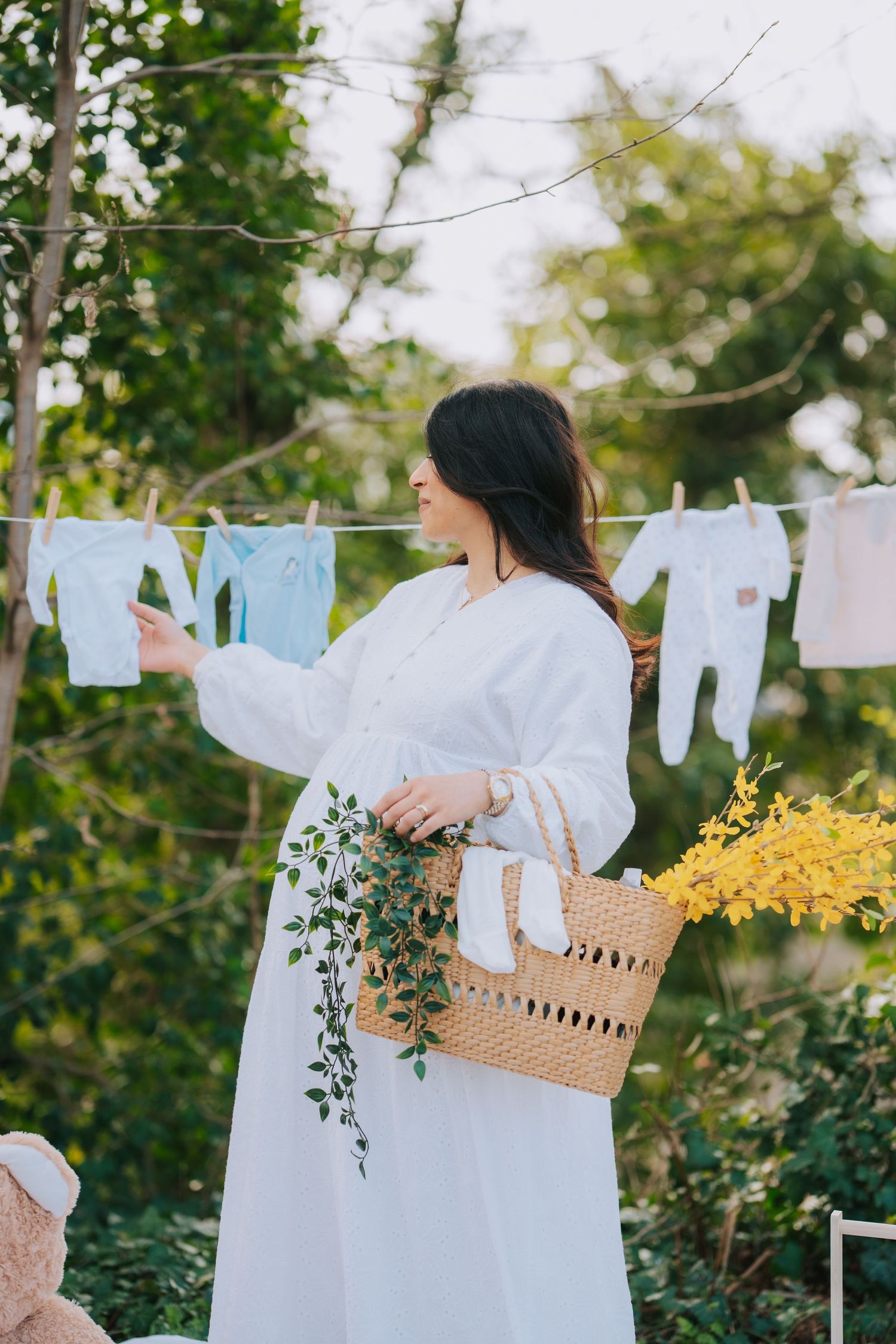 Person in flowing white dress hangs baby clothes on clothesline while holding woven basket with flowers in garden setting.