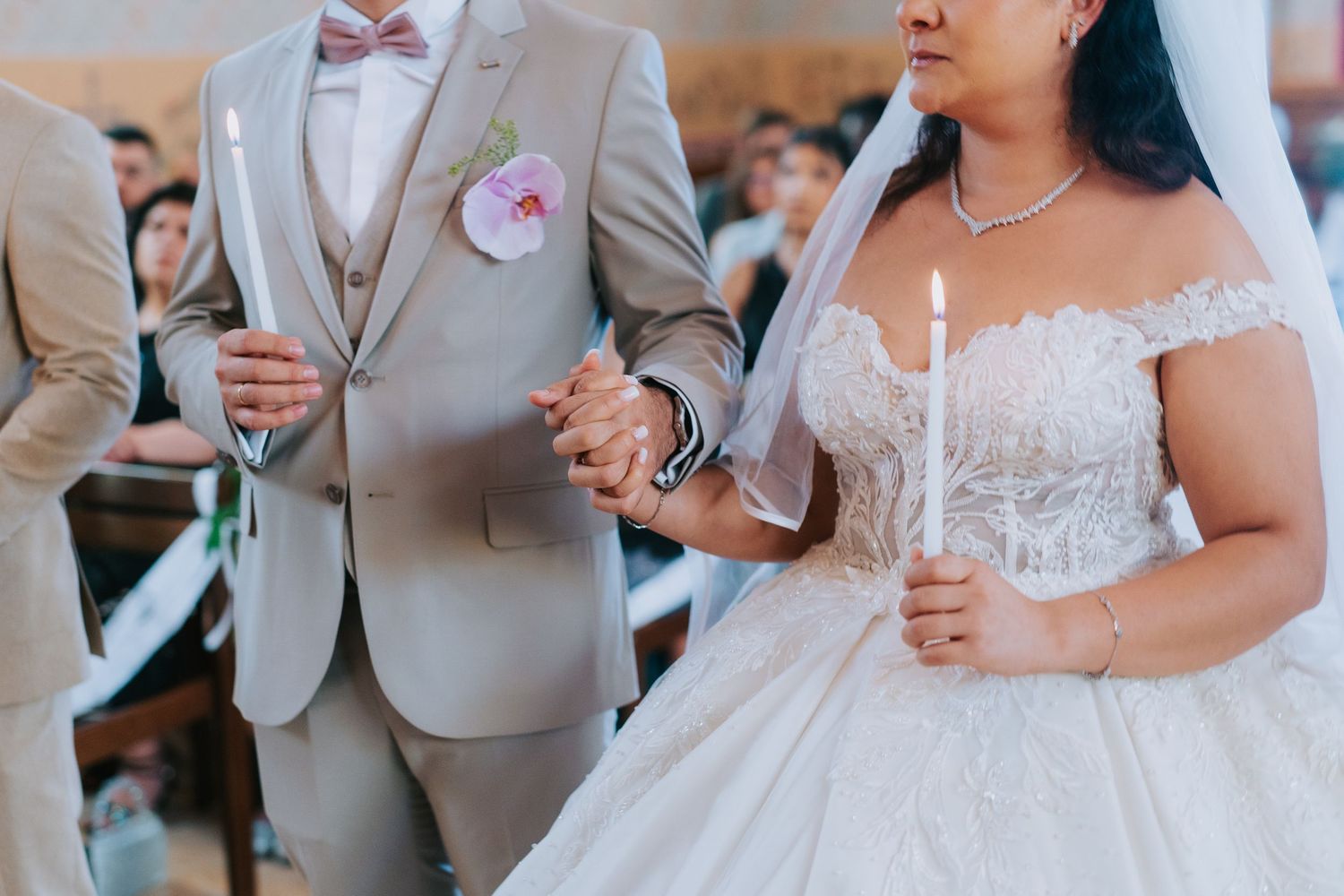 A couple holding hands at their wedding, with the groom in a light gray suit and the bride in a white lace gown.