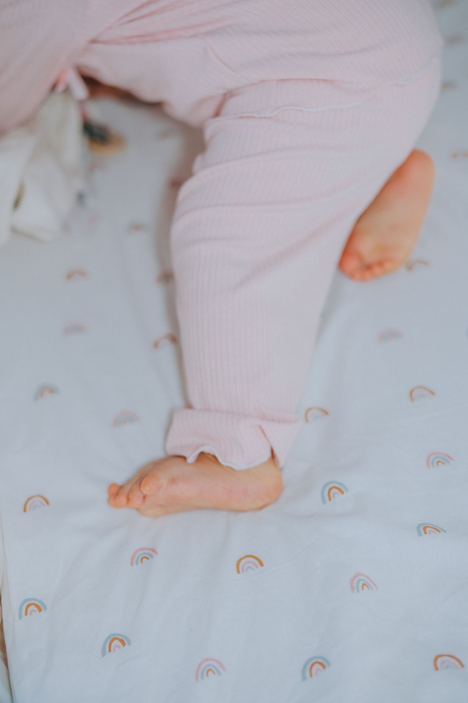Baby feet in pink pajamas on rainbow patterned bedding.