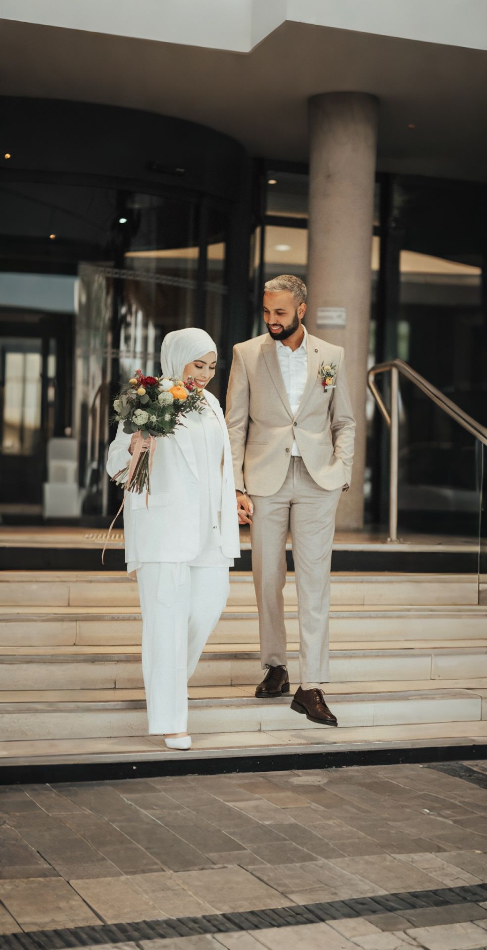 Newlyweds in matching light-colored attire descending modern building steps together.