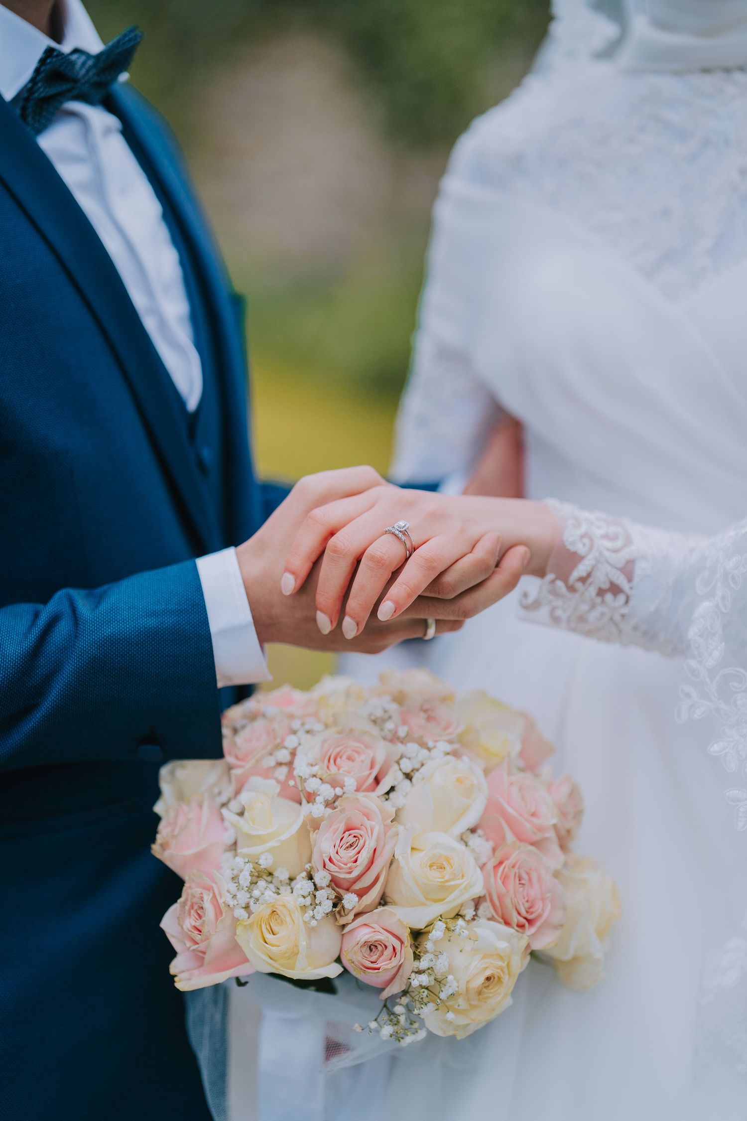 Couple holds hands with wedding rings while holding blush and ivory rose bridal bouquet against navy suit and white dress.