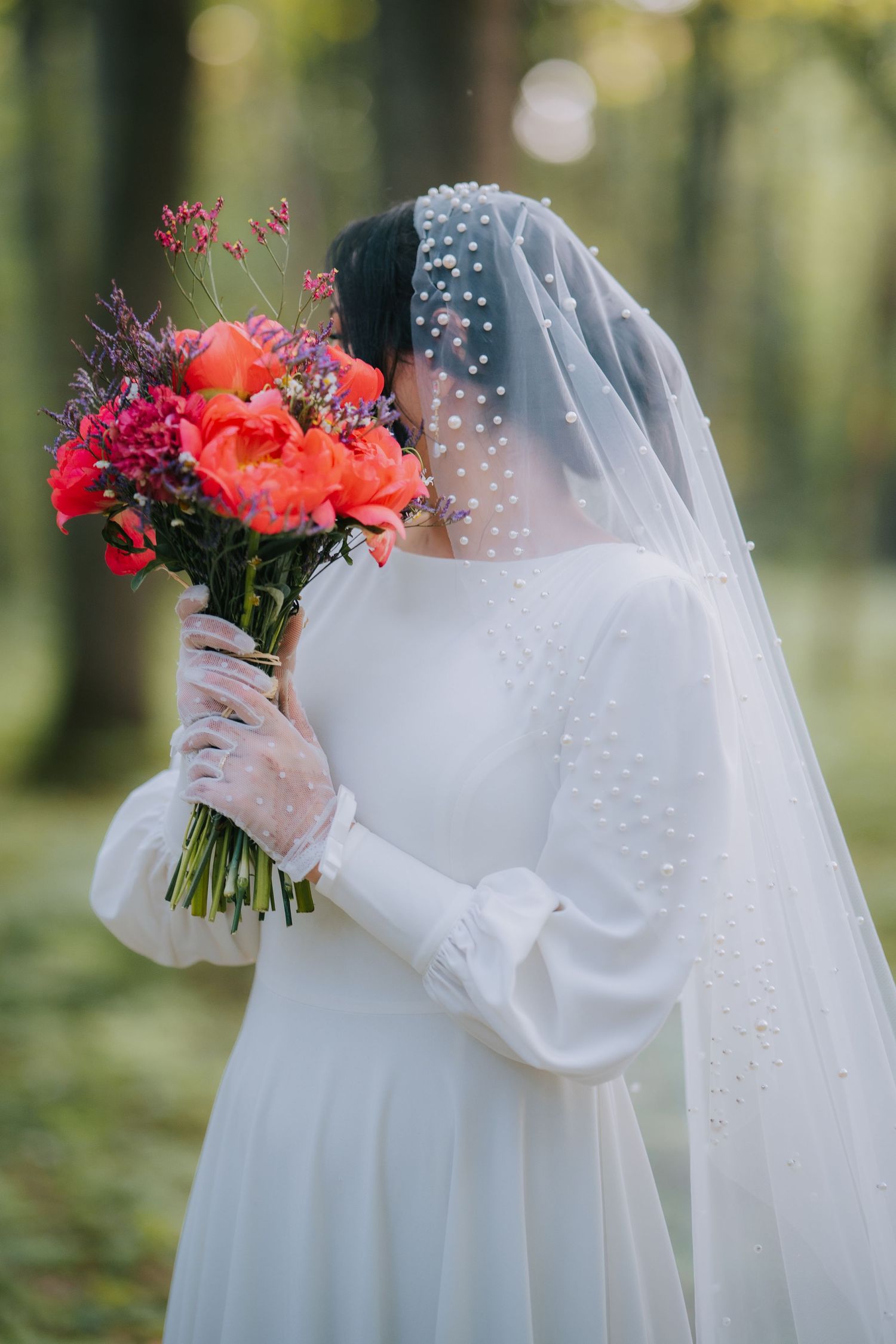 A bridal bouquet of vibrant coral roses and pink flowers held against a white wedding dress with pearl-studded veil.