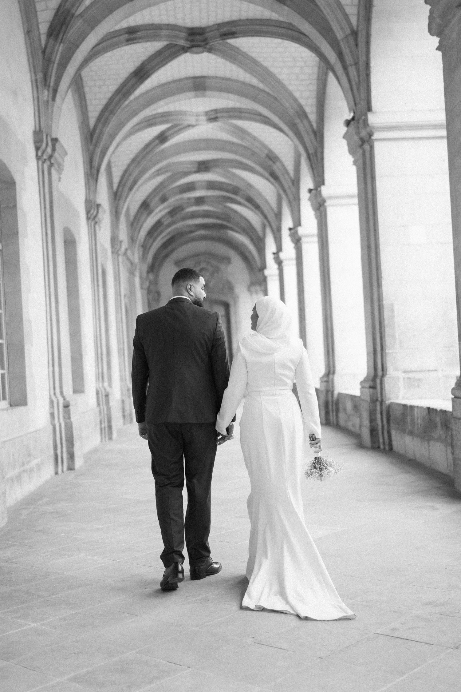 A couple in wedding attire walking down an elegant arched corridor in black and white photography.