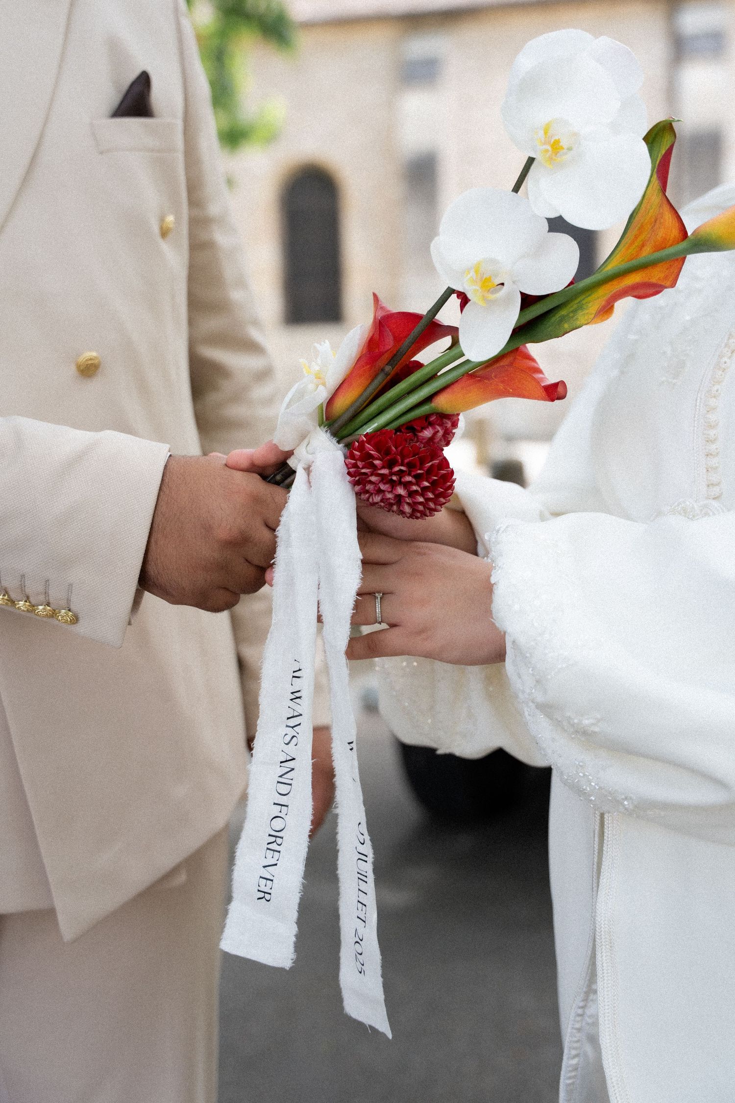 A wedding bouquet of white orchids and red calla lilies with decorative ribbons being exchanged between hands.
