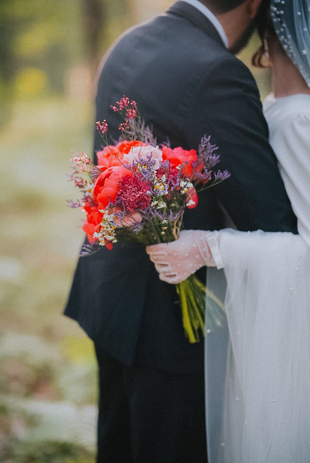Close-up of bride in white dress and groom in dark suit holding vibrant red and purple floral wedding bouquet.