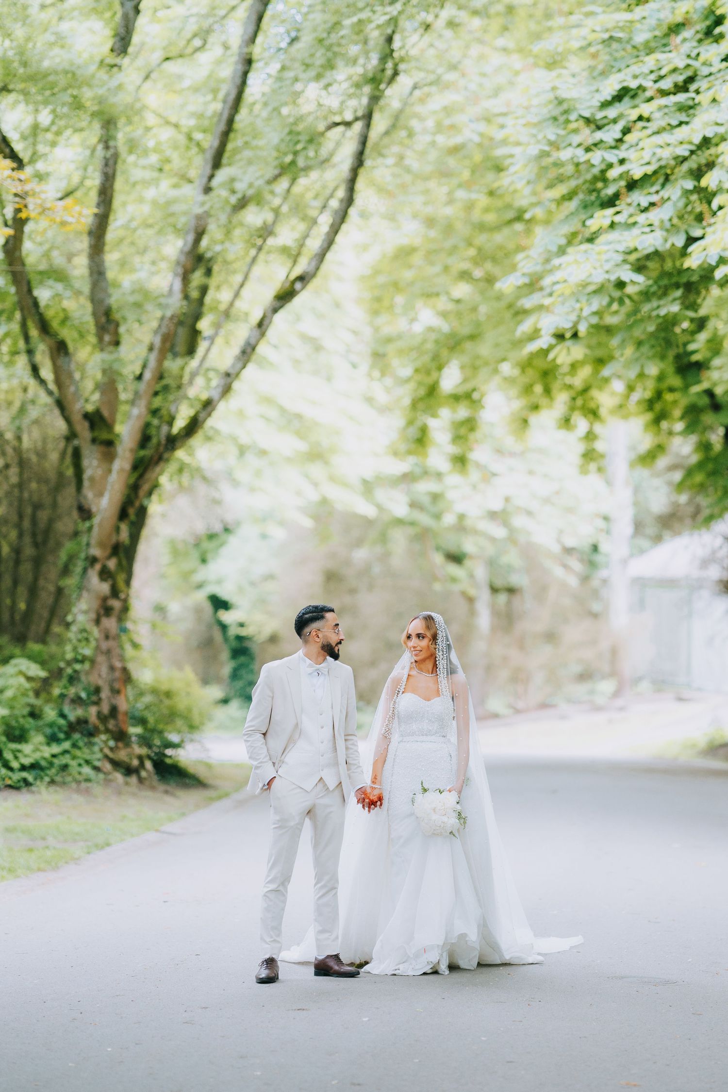 A couple in white wedding attire walk hand in hand down a tree-lined road on their special day.