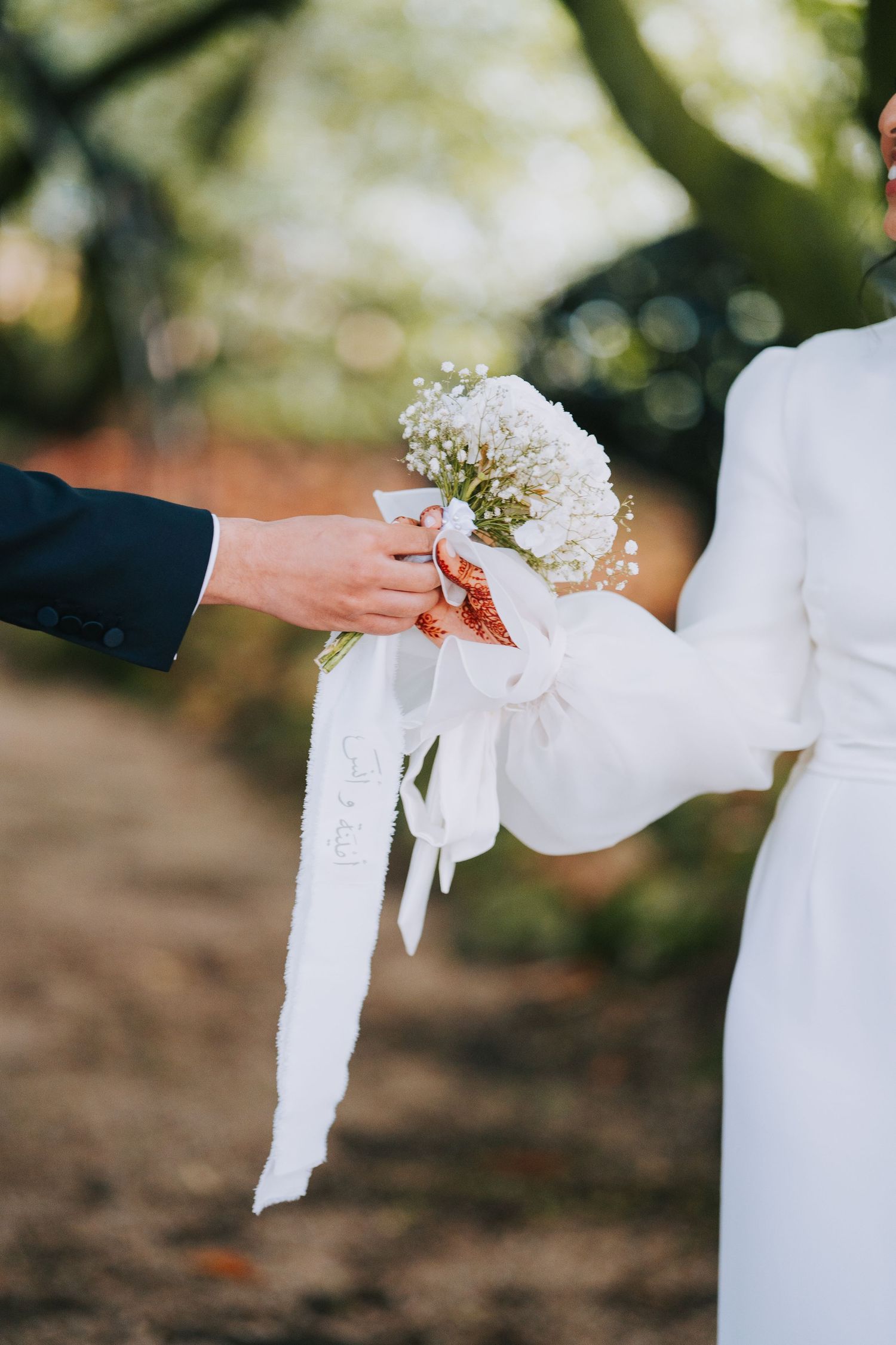 Two hands exchange a delicate white floral bouquet with flowing ribbons during an outdoor wedding ceremony.