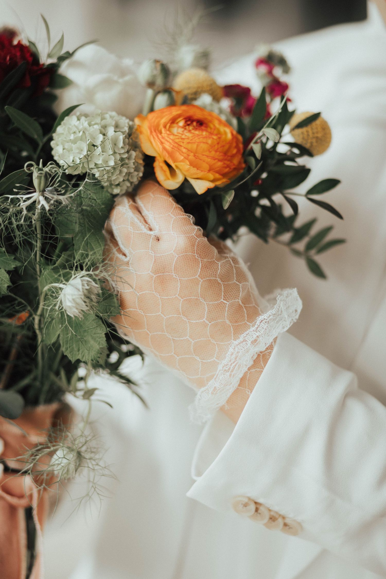 Close-up of delicate white lace and floral bouquet featuring orange ranunculus, hydrangeas and greenery.
