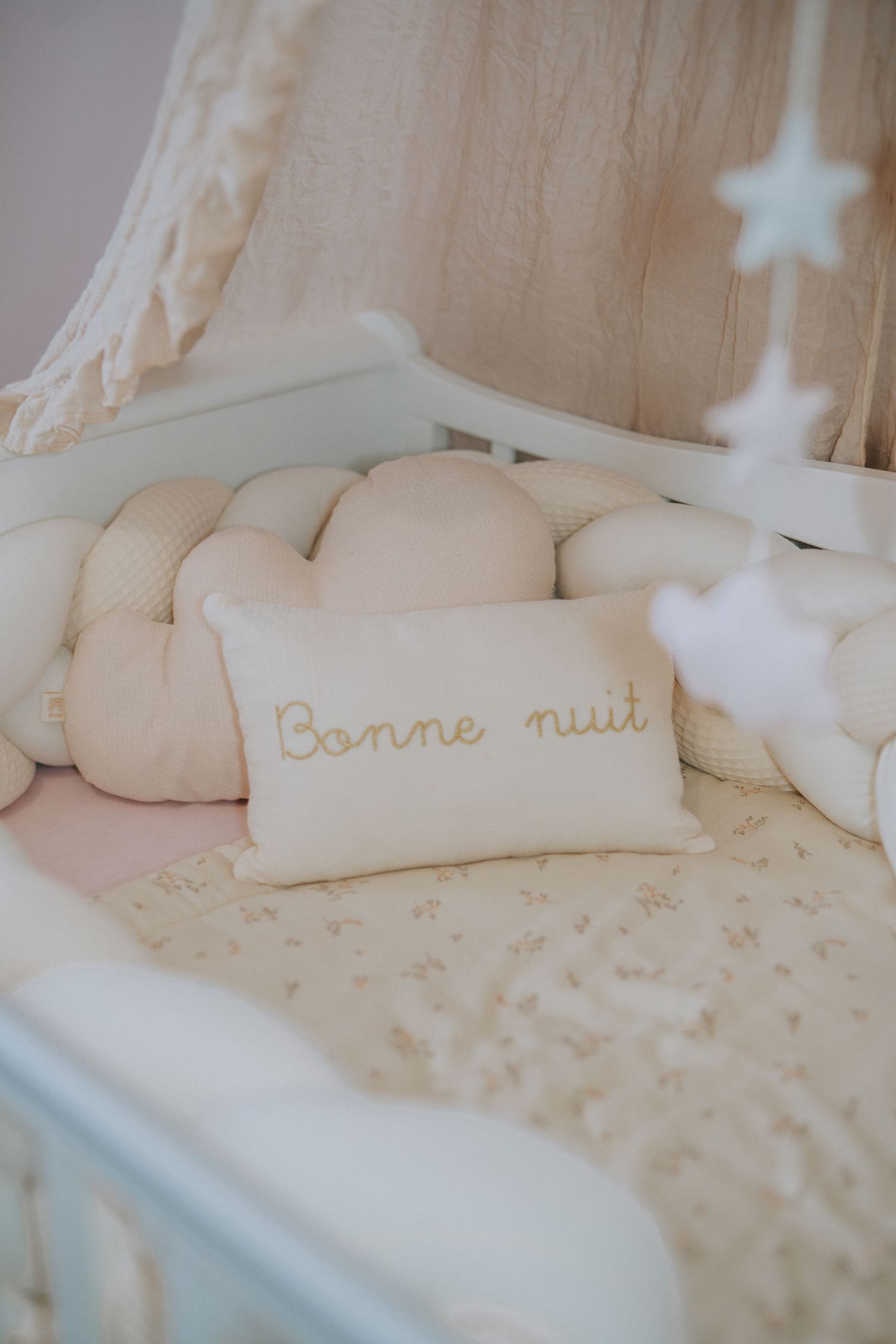 White crib with plush cushions and 'Bonne nuit' pillow under hanging stars.