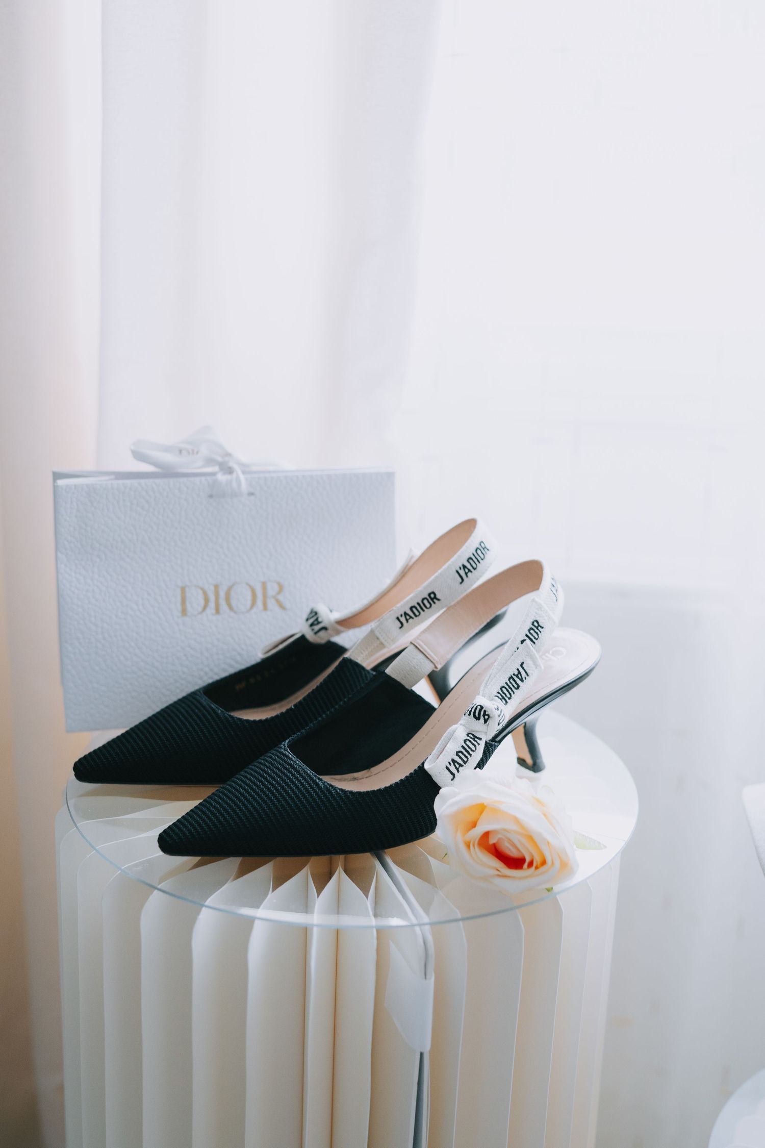 A pair of black Dior slingback heels with mesh detail displayed on a white sculptural pedestal next to a shopping bag and orchid.