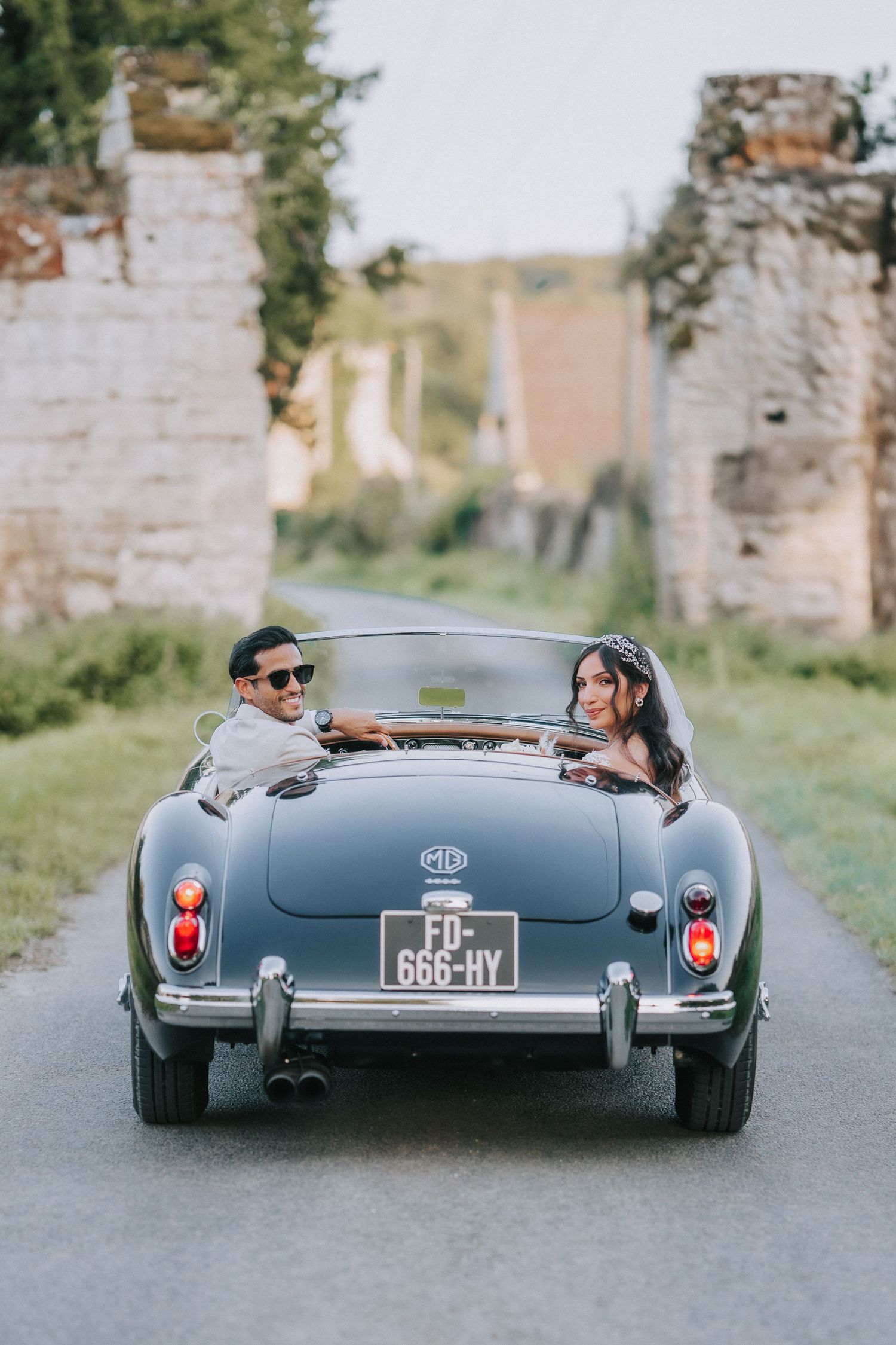 Classic MG convertible sports car driving away on a country road past stone ruins in a romantic setting.