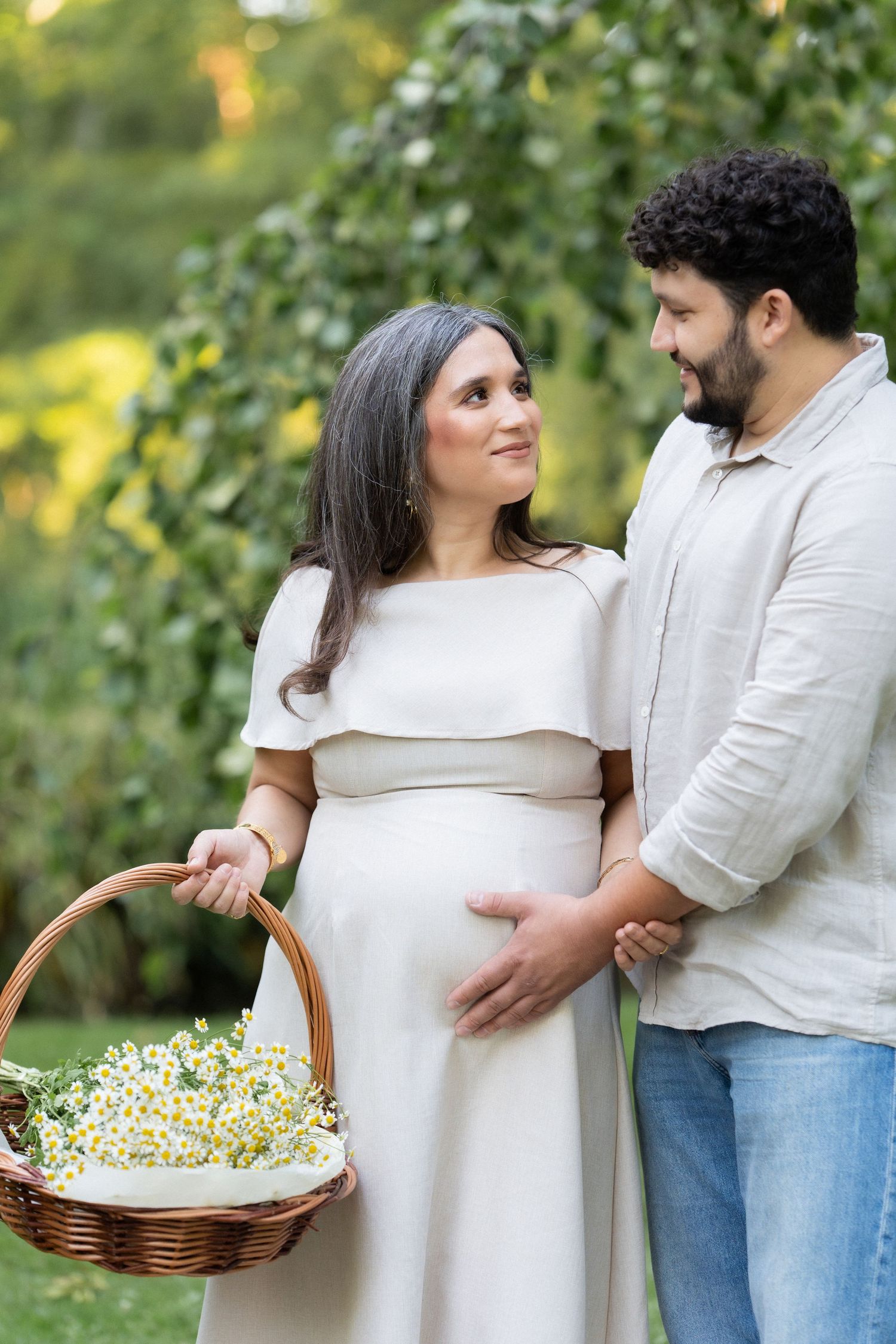 Expectant couple in white outfits share a tender moment outdoors while holding a basket of daisies in a garden setting.