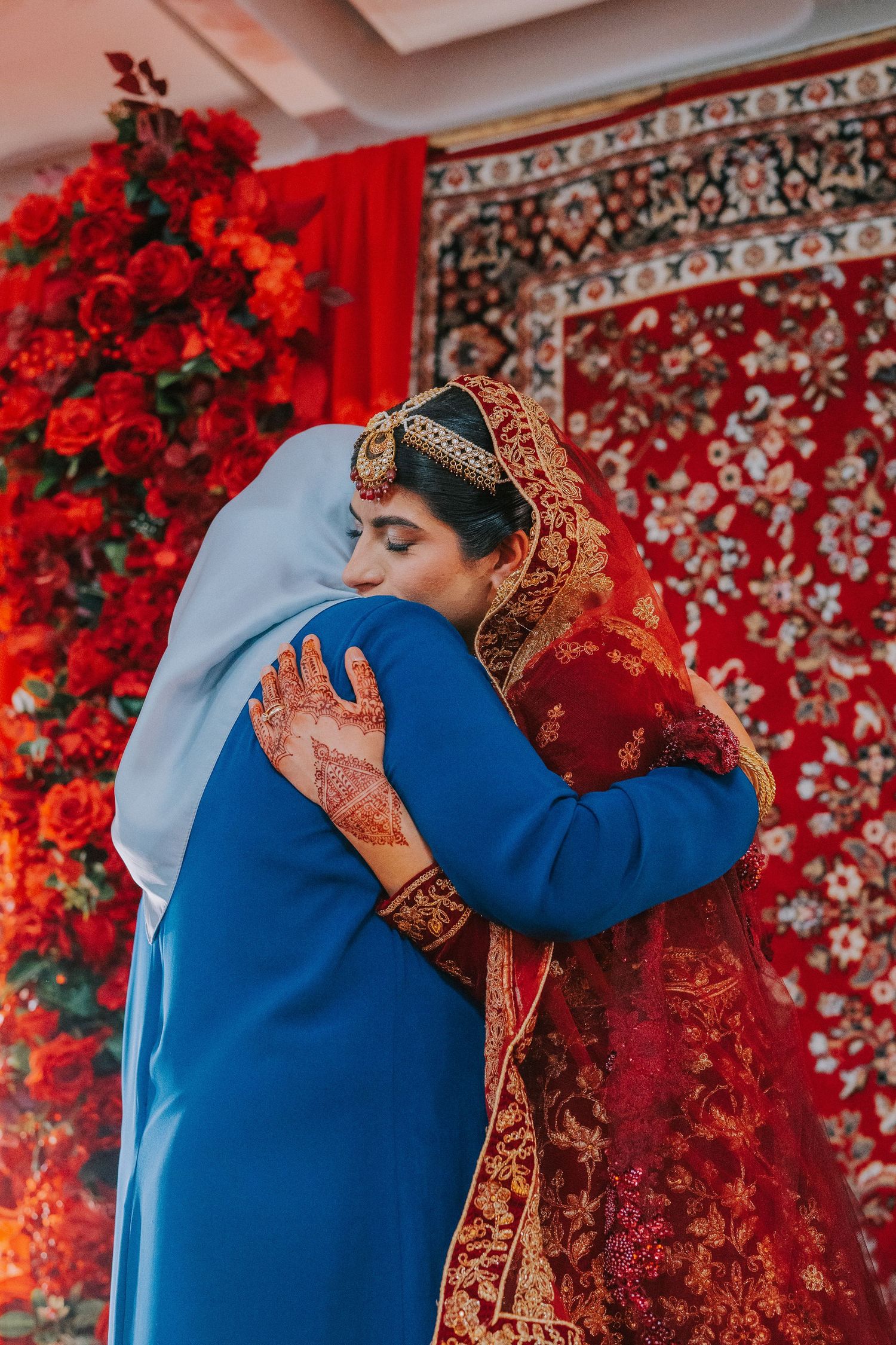 A traditional South Asian wedding embrace against a vibrant red floral backdrop and patterned tapestry.