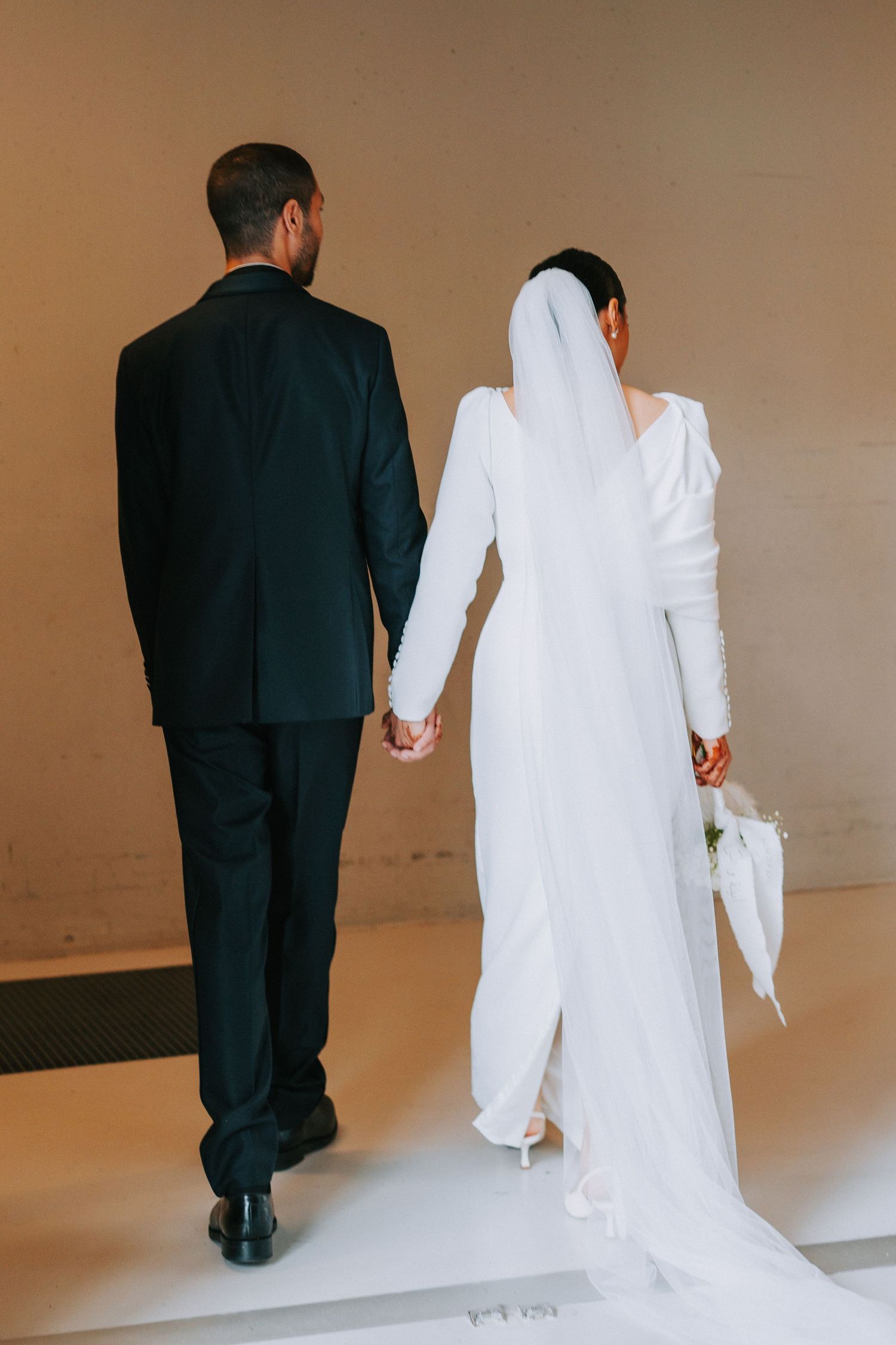 Newlywed couple in black suit and white dress holding hands while walking away from camera on light brown background.