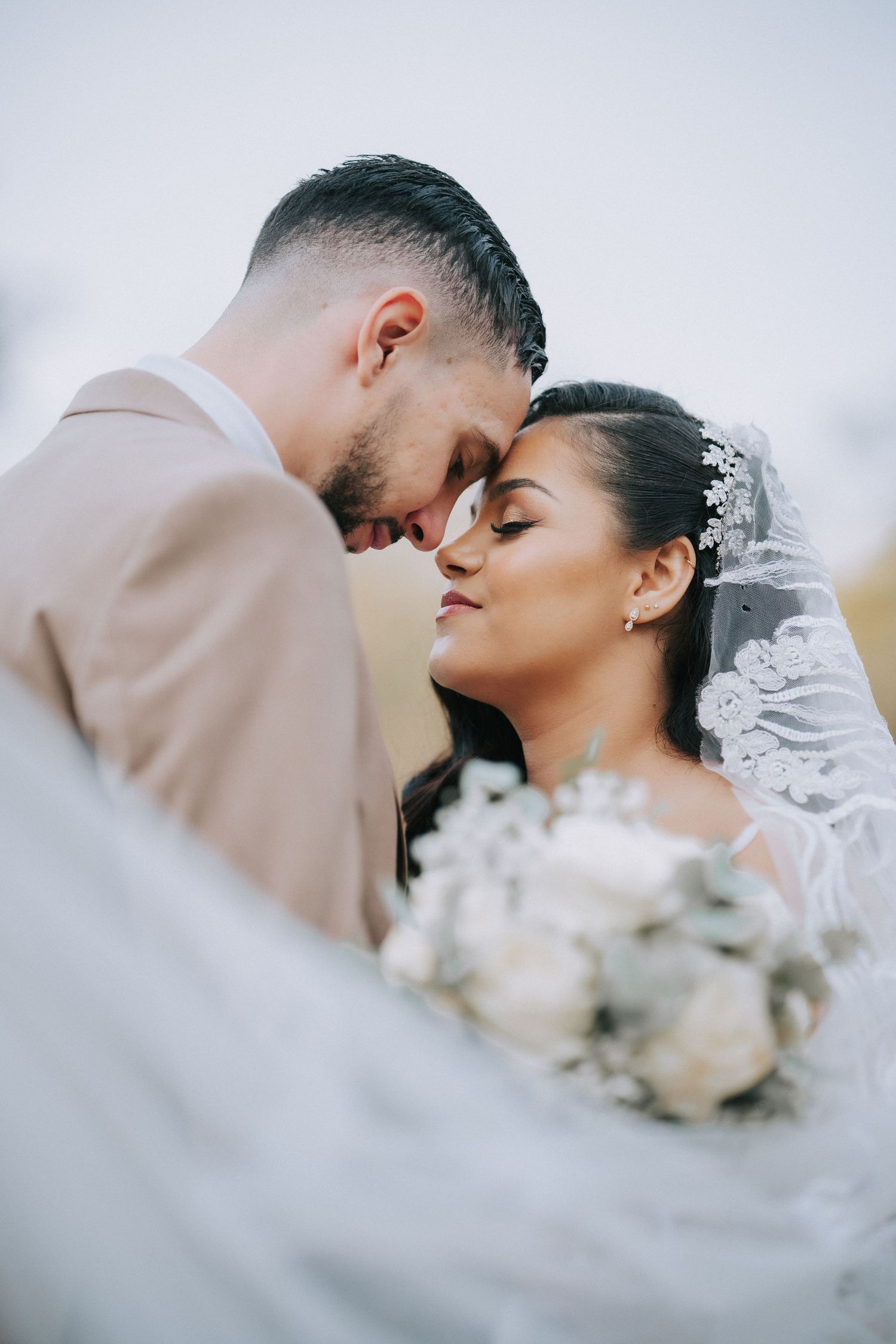 Tender wedding moment captured as couple shares intimate foreheads-touching pose with bridal bouquet and veil details.