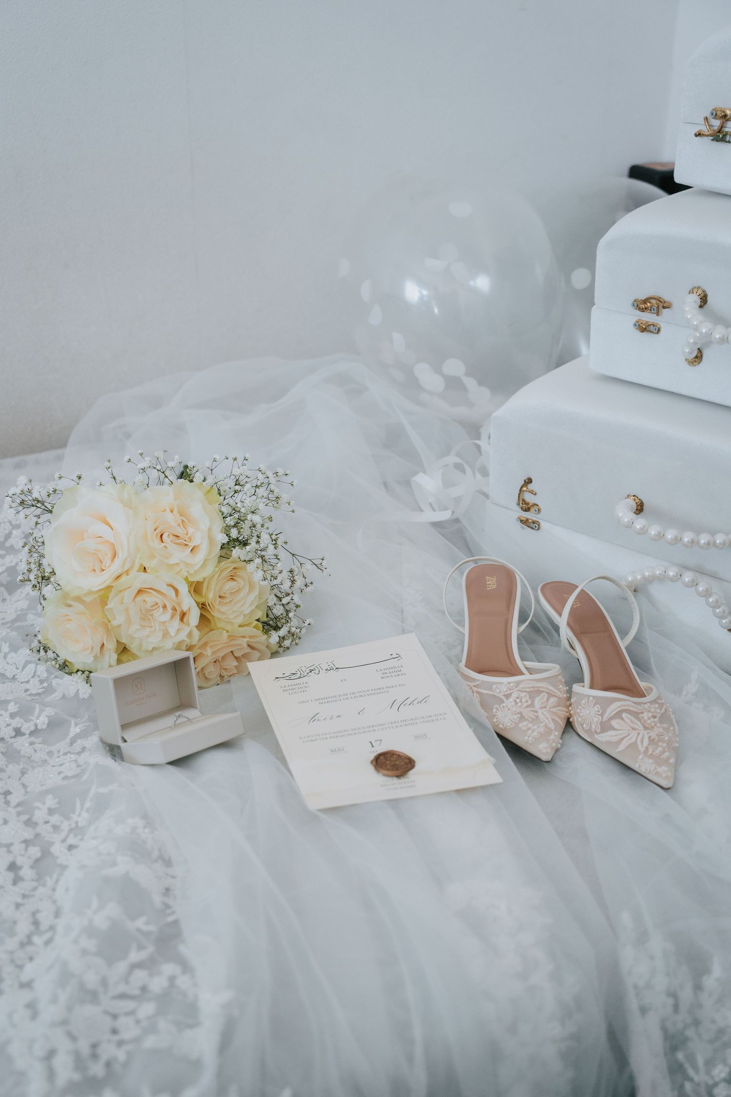 Wedding details including white tulle dress, blush heels, delicate invitation and soft peach floral arrangements on white backdrop.