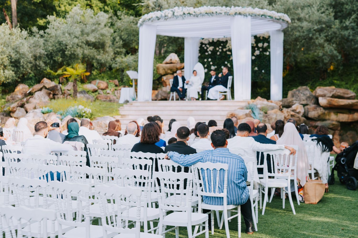Outdoor wedding ceremony setup with white chairs and a decorated gazebo altar in a garden setting.