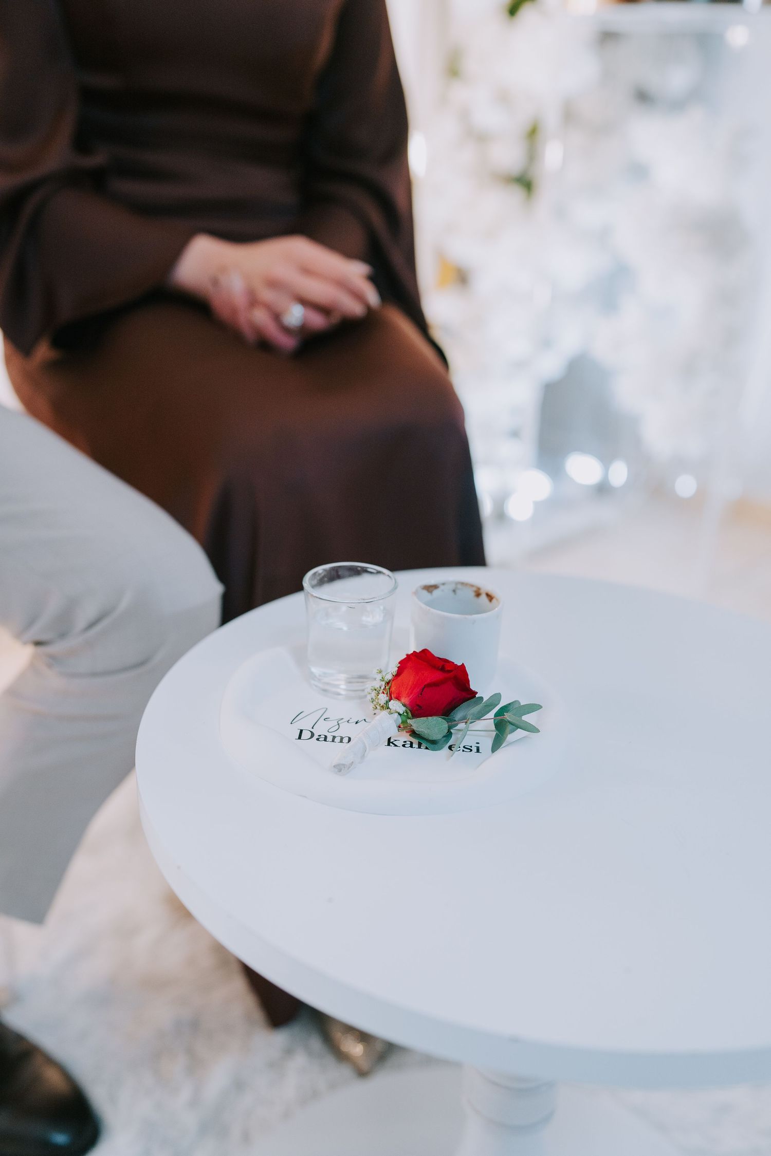 A single red rose and glass sit on a white round table with someone sitting nearby in brown attire.