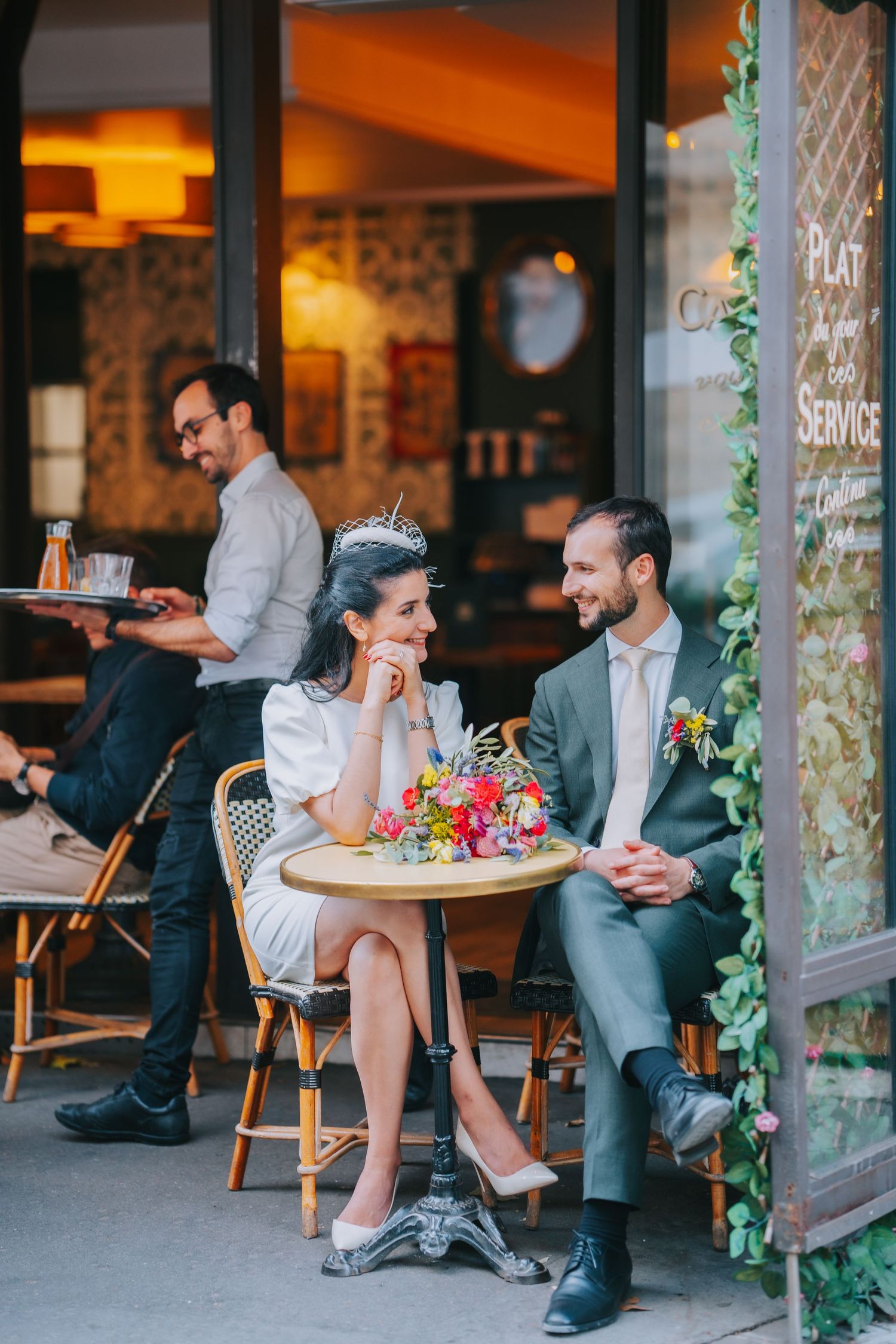 Couple dining at romantic Parisian style cafe with waiter serving in background and decorative ivy on storefront.