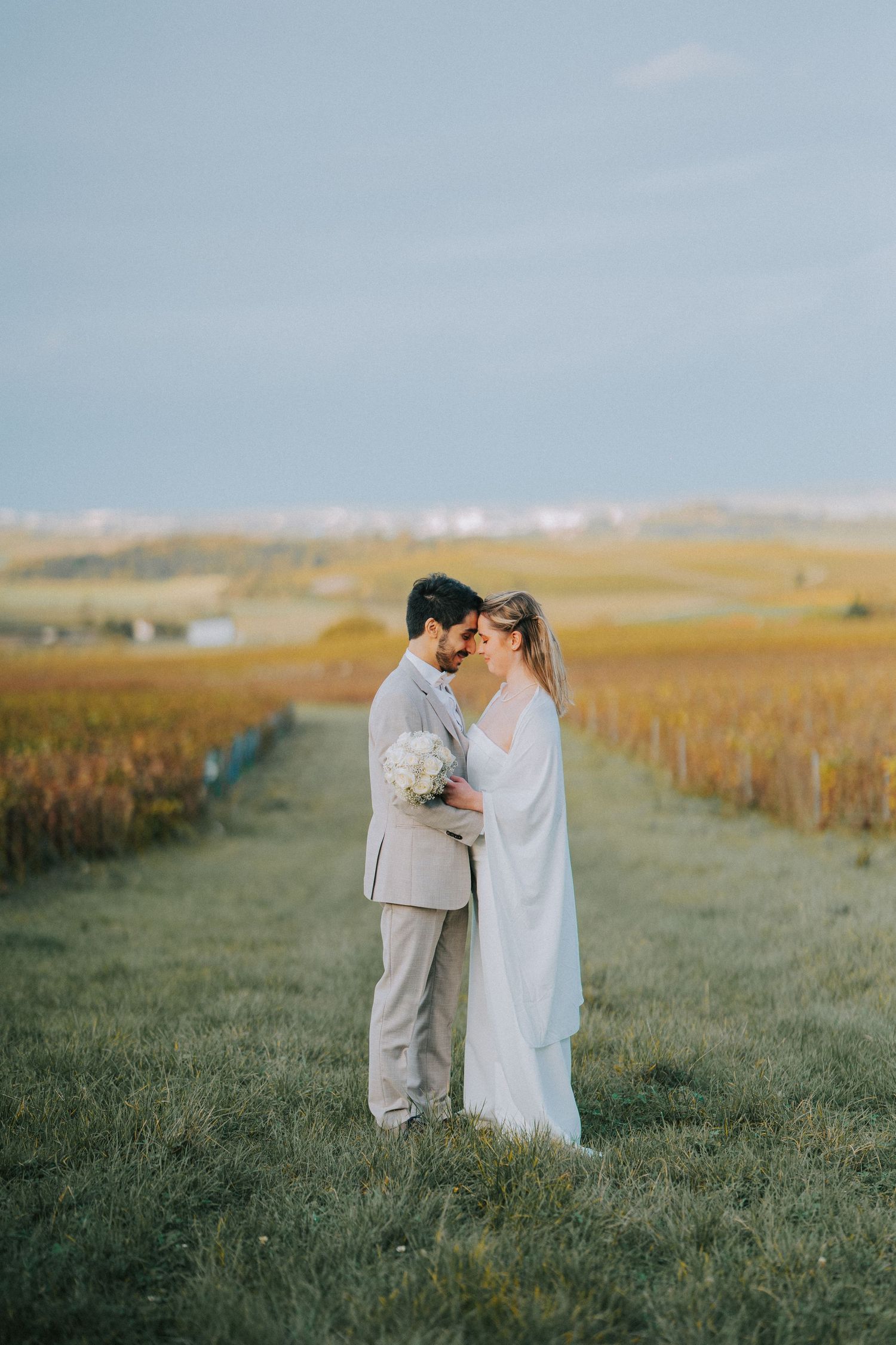 Two newlyweds share an intimate moment on a grassy path between golden wheat fields at sunset.