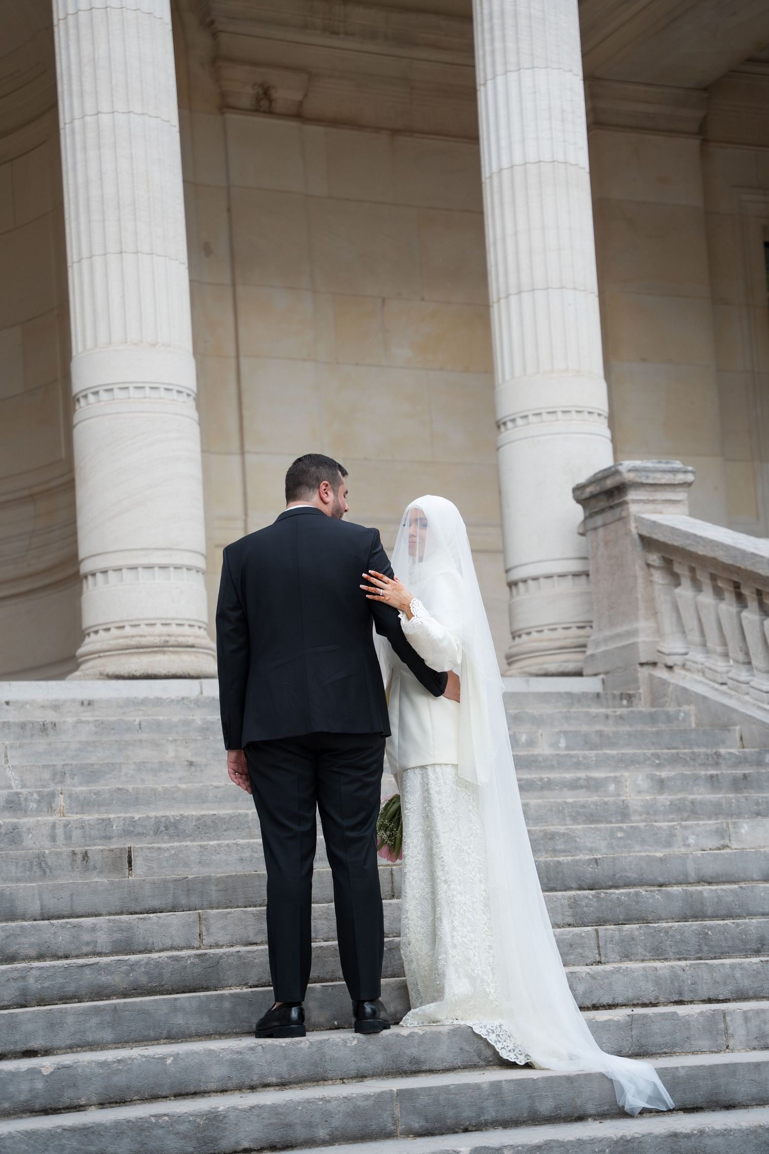 A couple in wedding attire shares an intimate moment on grand stone steps with white columns in the background.