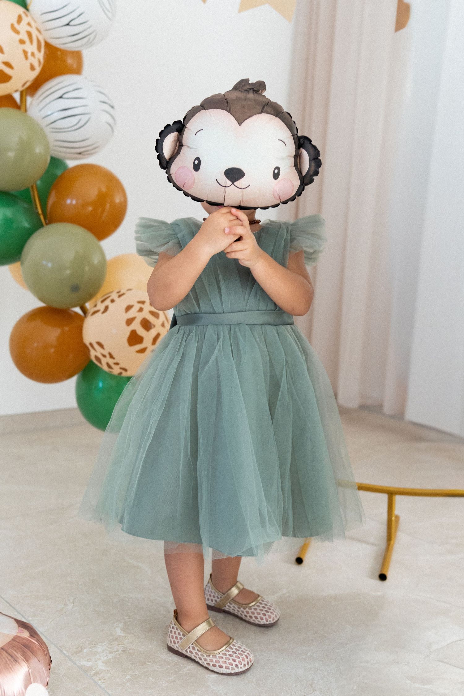 A child in a sage green tulle dress and polka dot flats twirls among party balloons and decorations.