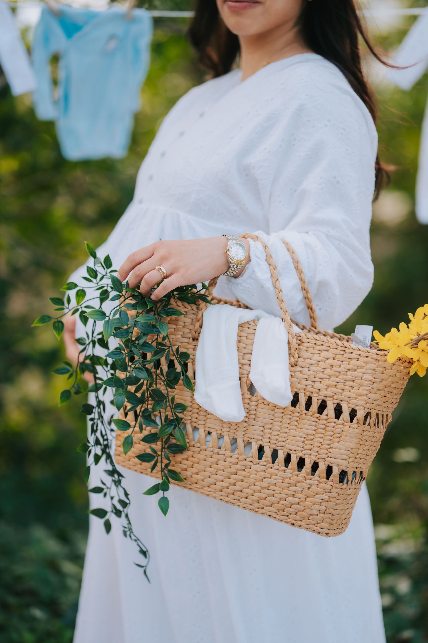 Woven straw handbag with green plants and flowers against white dress in outdoor setting.