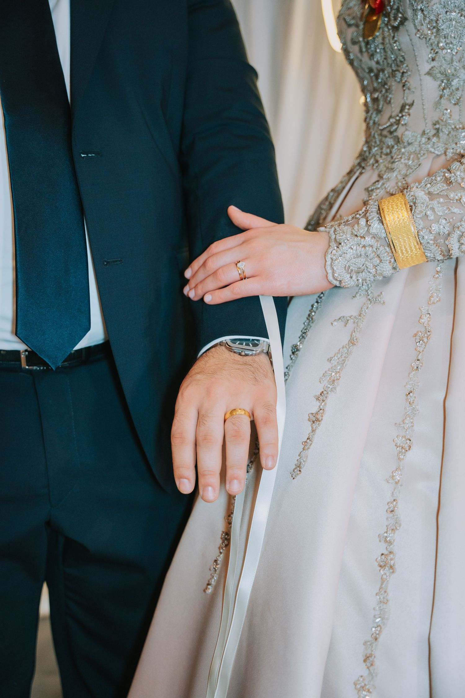 Close-up of bride and groom's hands showing wedding rings and formal attire.