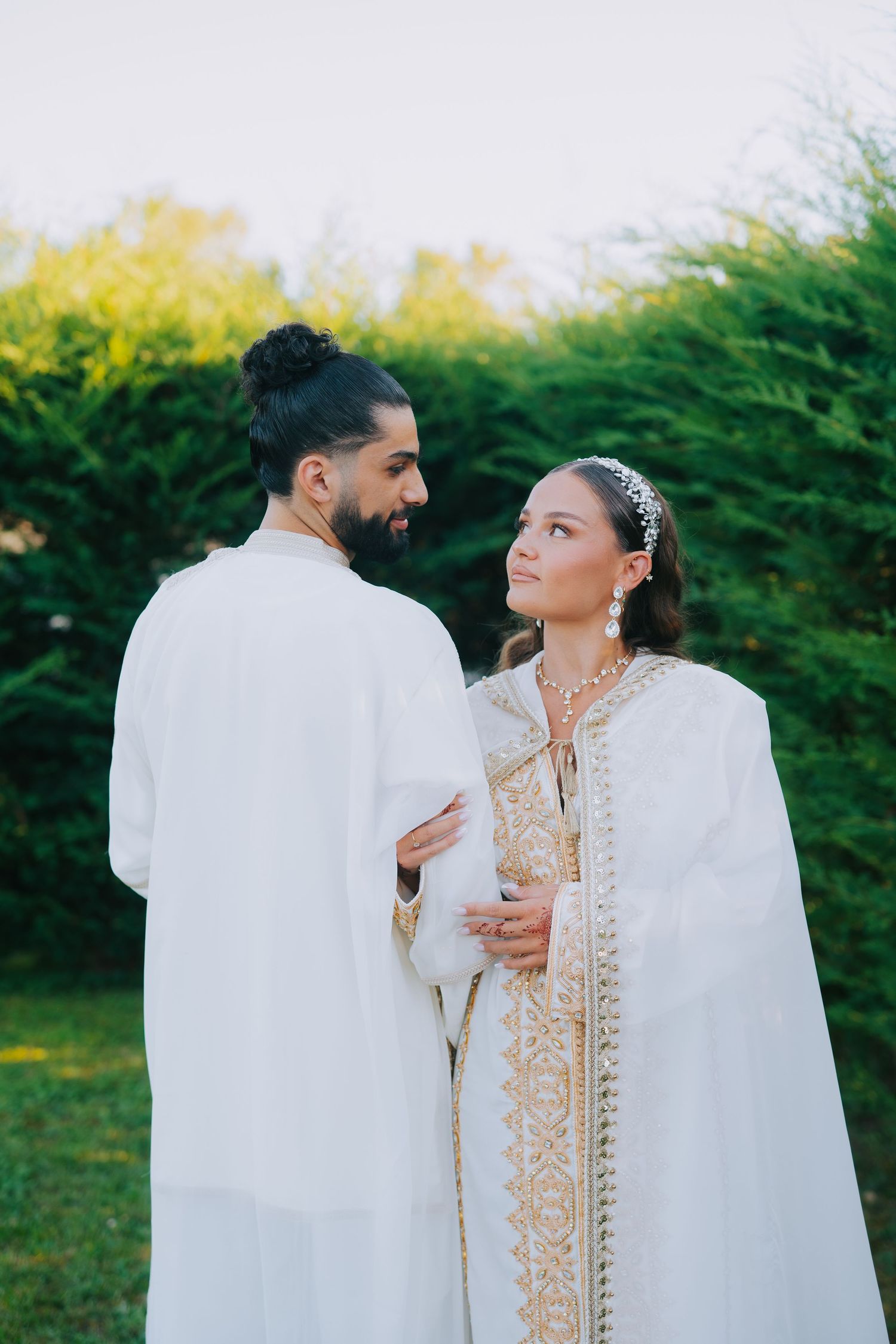 Couple wearing elegant white traditional Moroccan wedding attire standing in a garden setting.