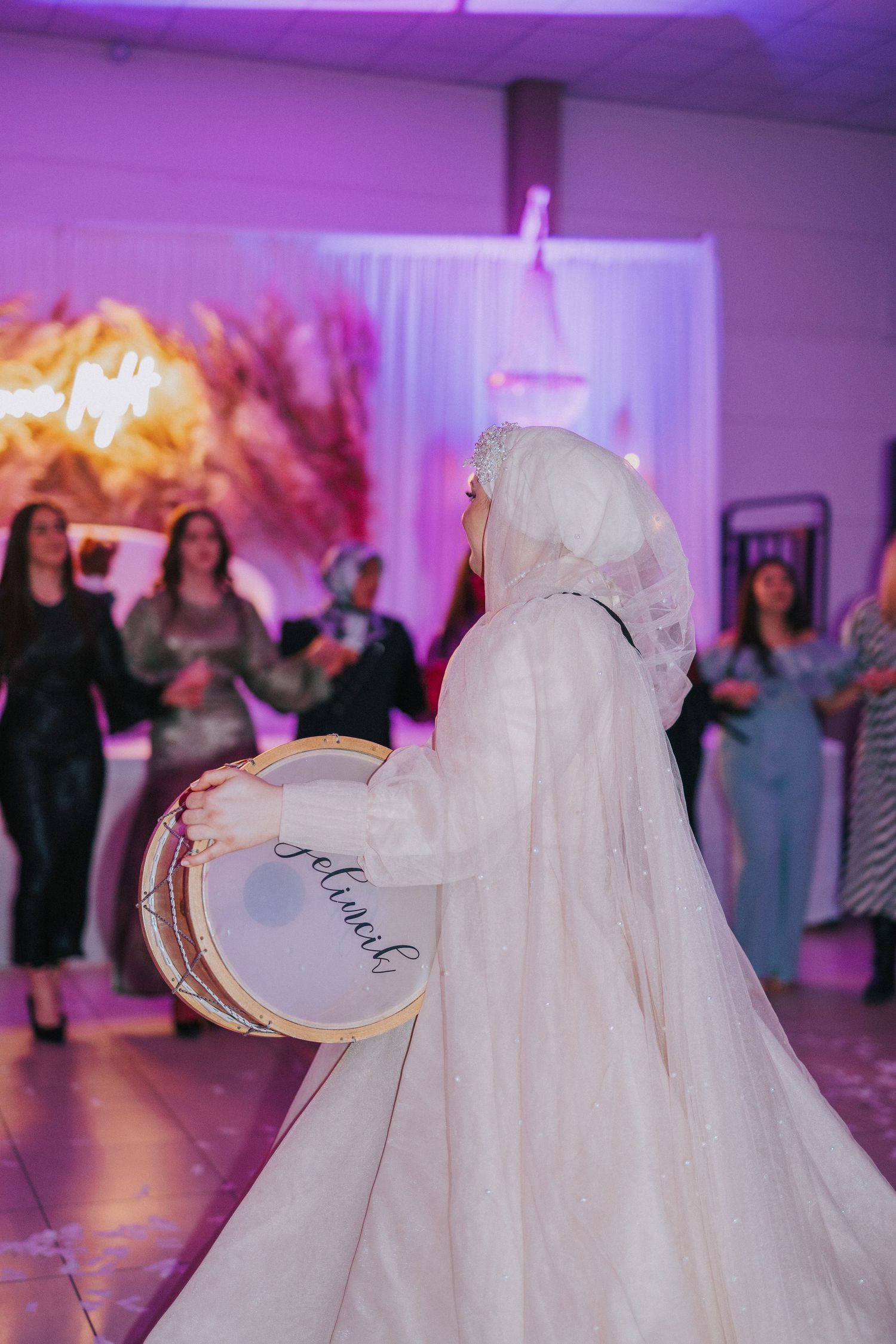 Person in flowing white dress holding decorative drum during wedding celebration.