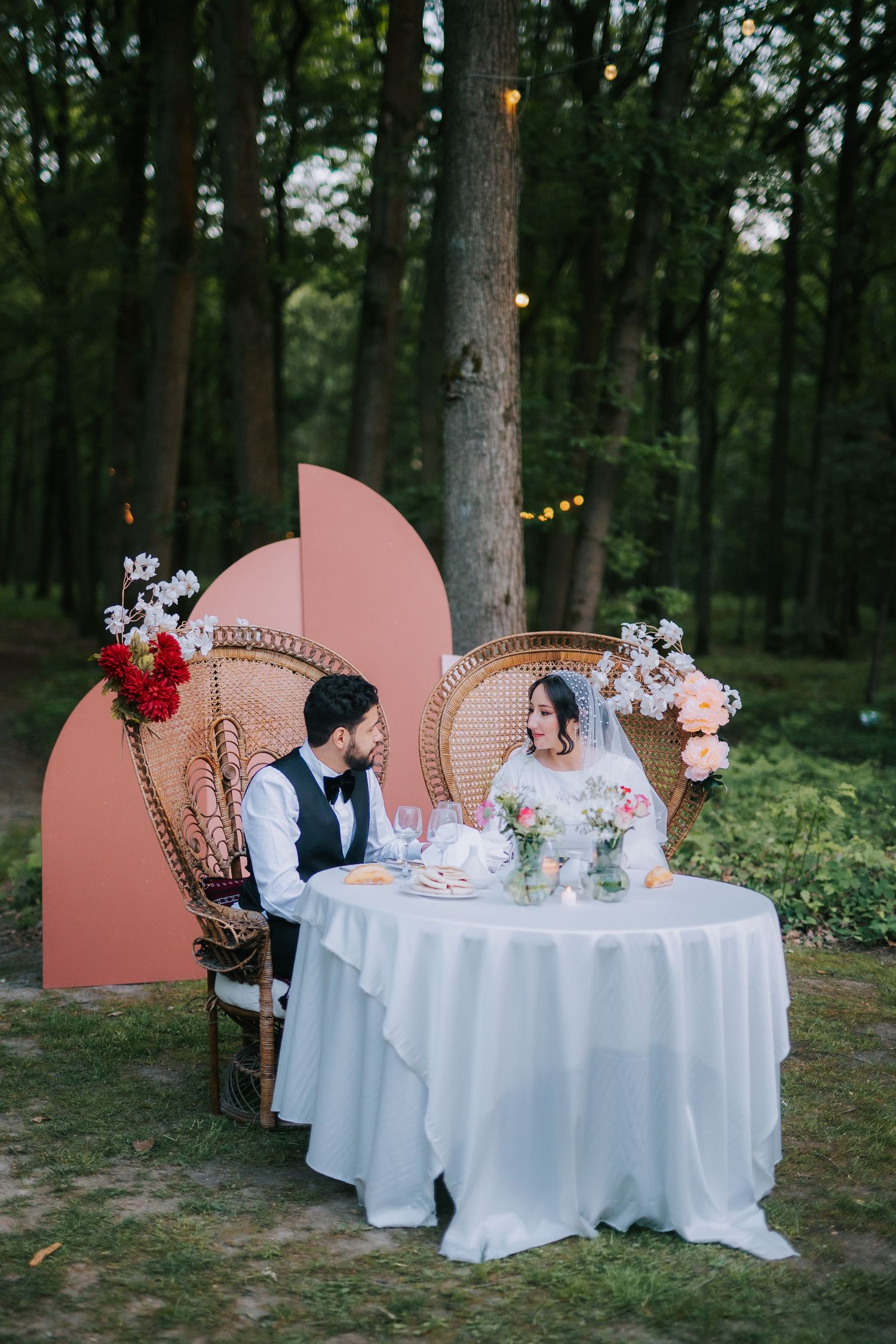 An intimate wedding reception table setup in a forest with pink decorative panels, string lights and floral arrangements.