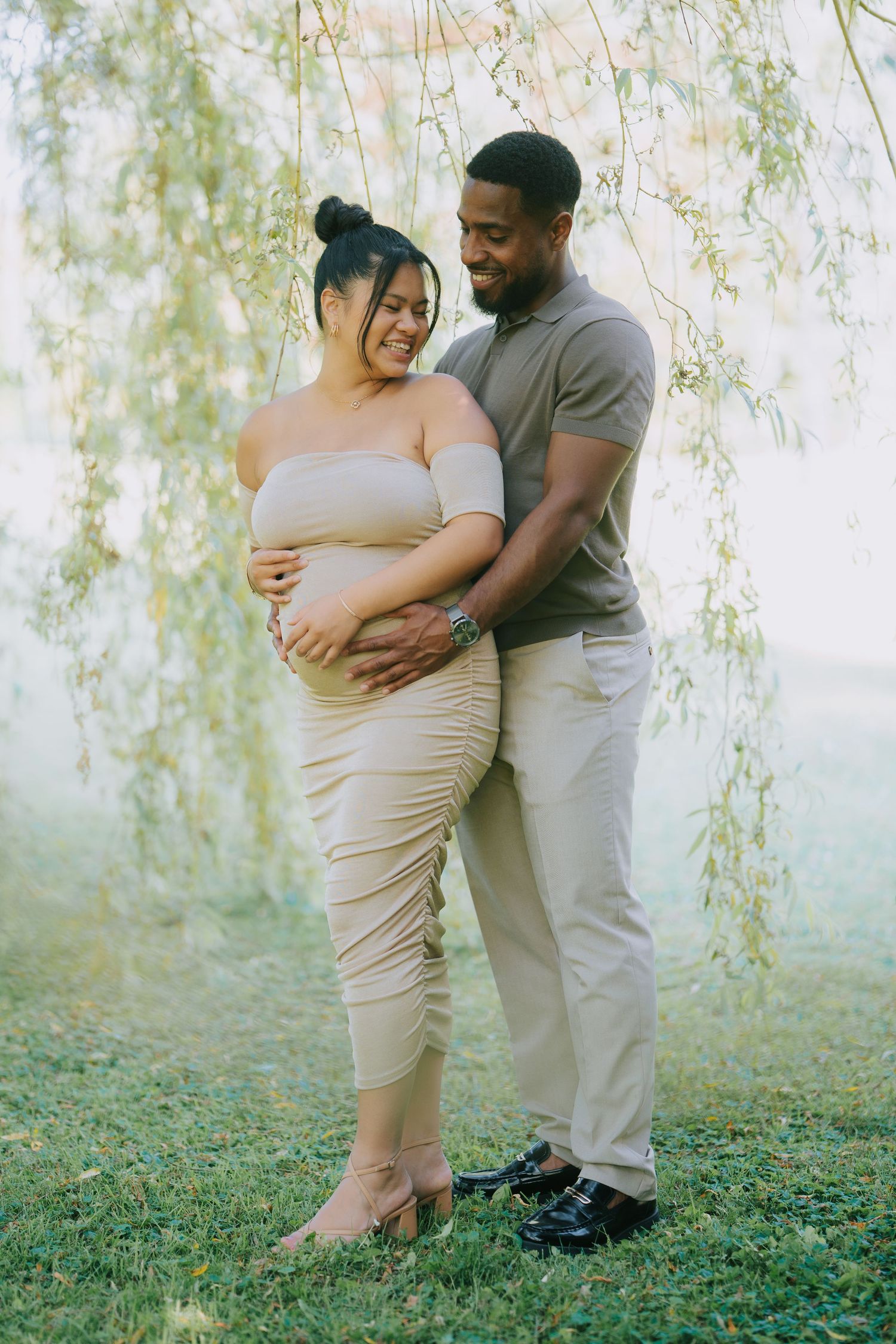 Expecting couple embraces under willow tree branches in dreamy maternity photo session at sunset.