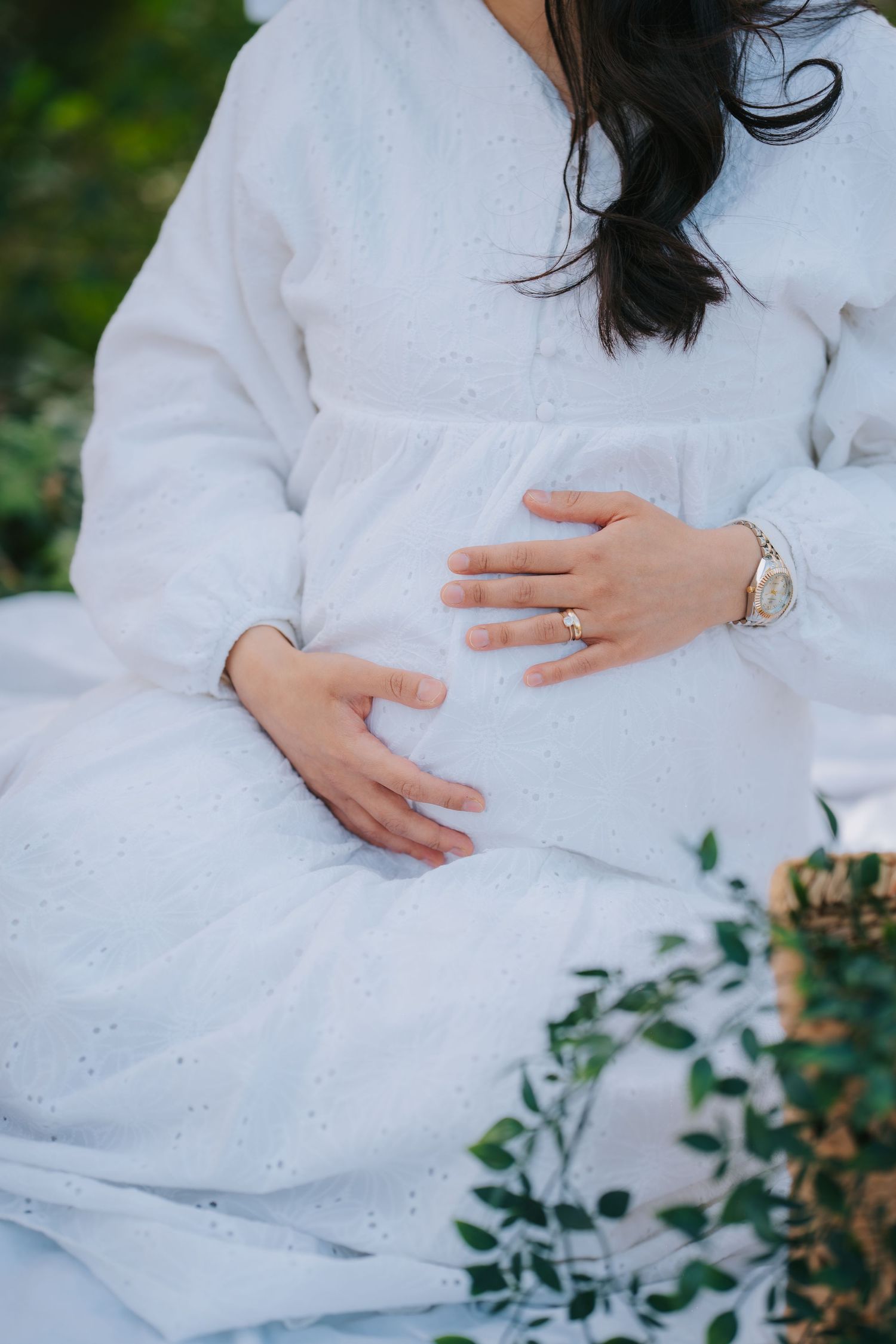 Close-up of hands cradling pregnant belly in white dress with engagement ring visible.