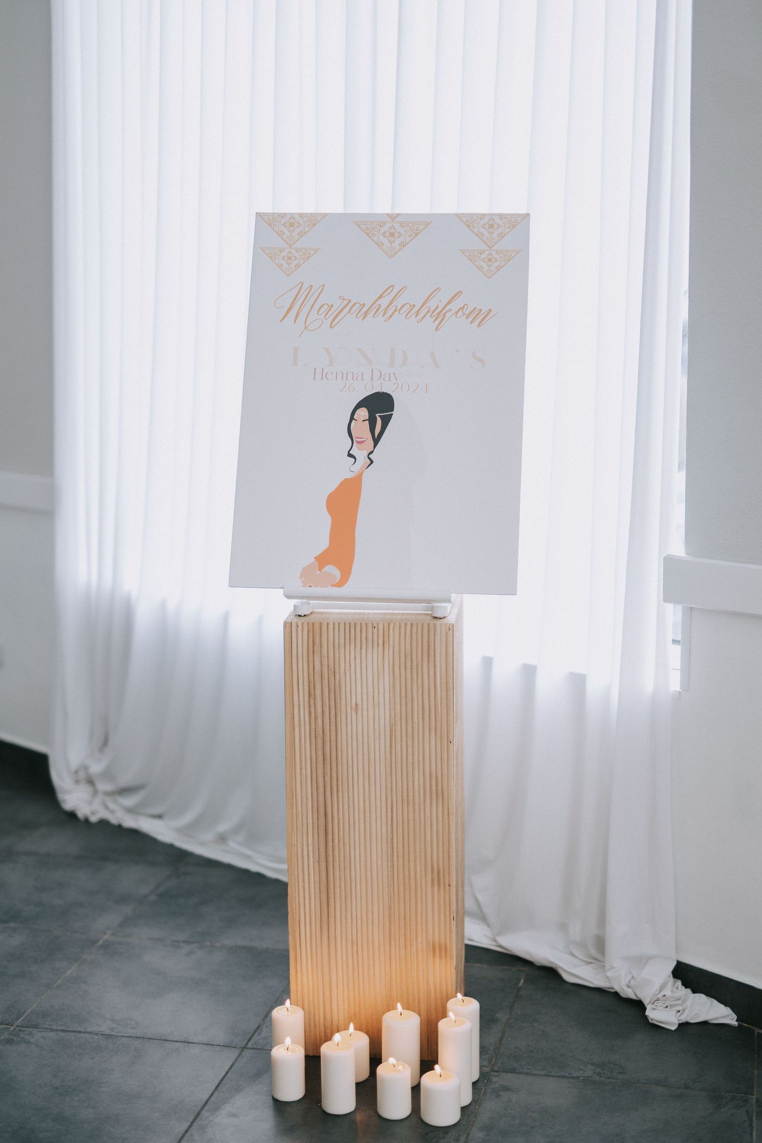 Wedding welcome sign on wooden pedestal surrounded by white candles against draped backdrop.