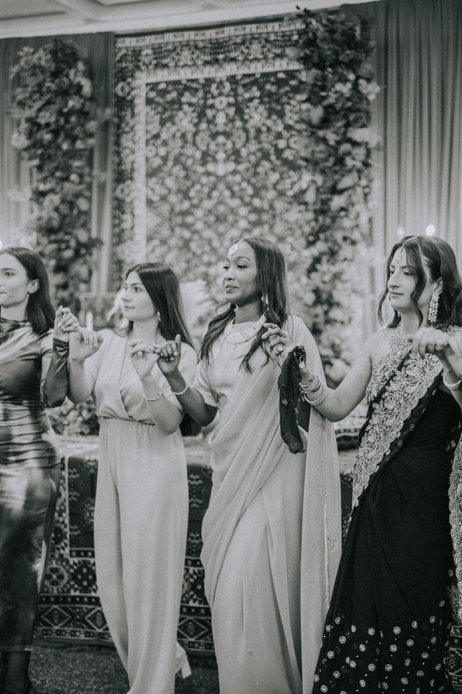 Group of guests wearing elegant gowns dancing together at a formal celebration with floral decor in background.