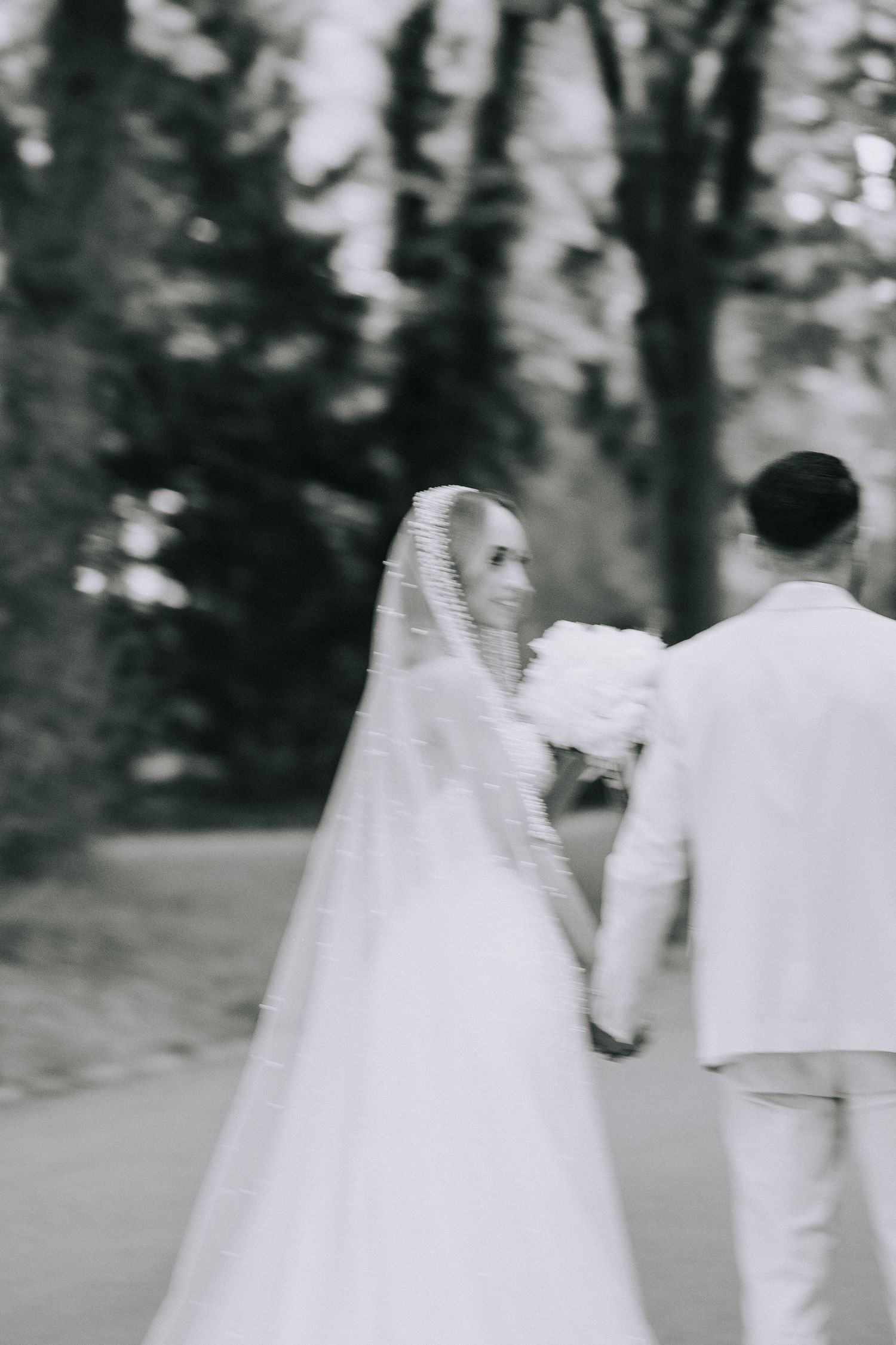 Black and white photo of a bride in a flowing white gown and veil walking with someone in white on a garden path.