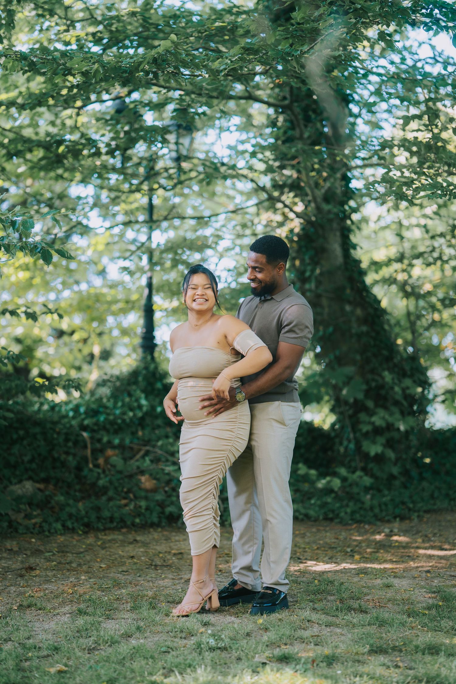 A couple embraces outdoors in a sunlit wooded setting, with the person in beige standing barefoot on grass.