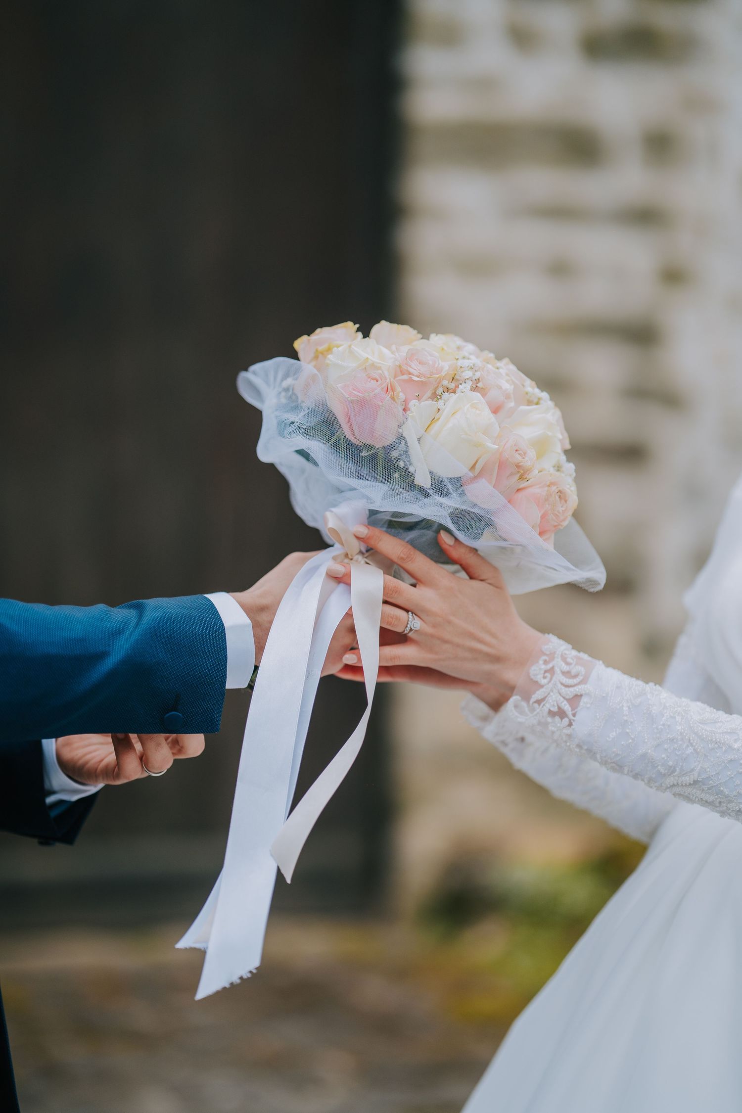 Close-up of hands exchanging a delicate bridal bouquet with blush pink flowers and white ribbons during a wedding ceremony.