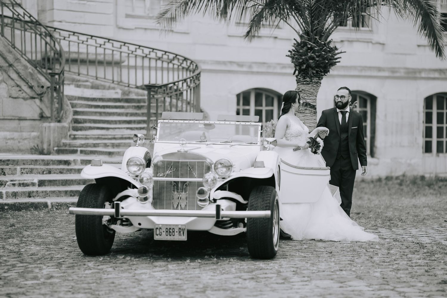 Vintage black and white photograph showing a classic car parked in front of an elegant mansion with curved staircase.