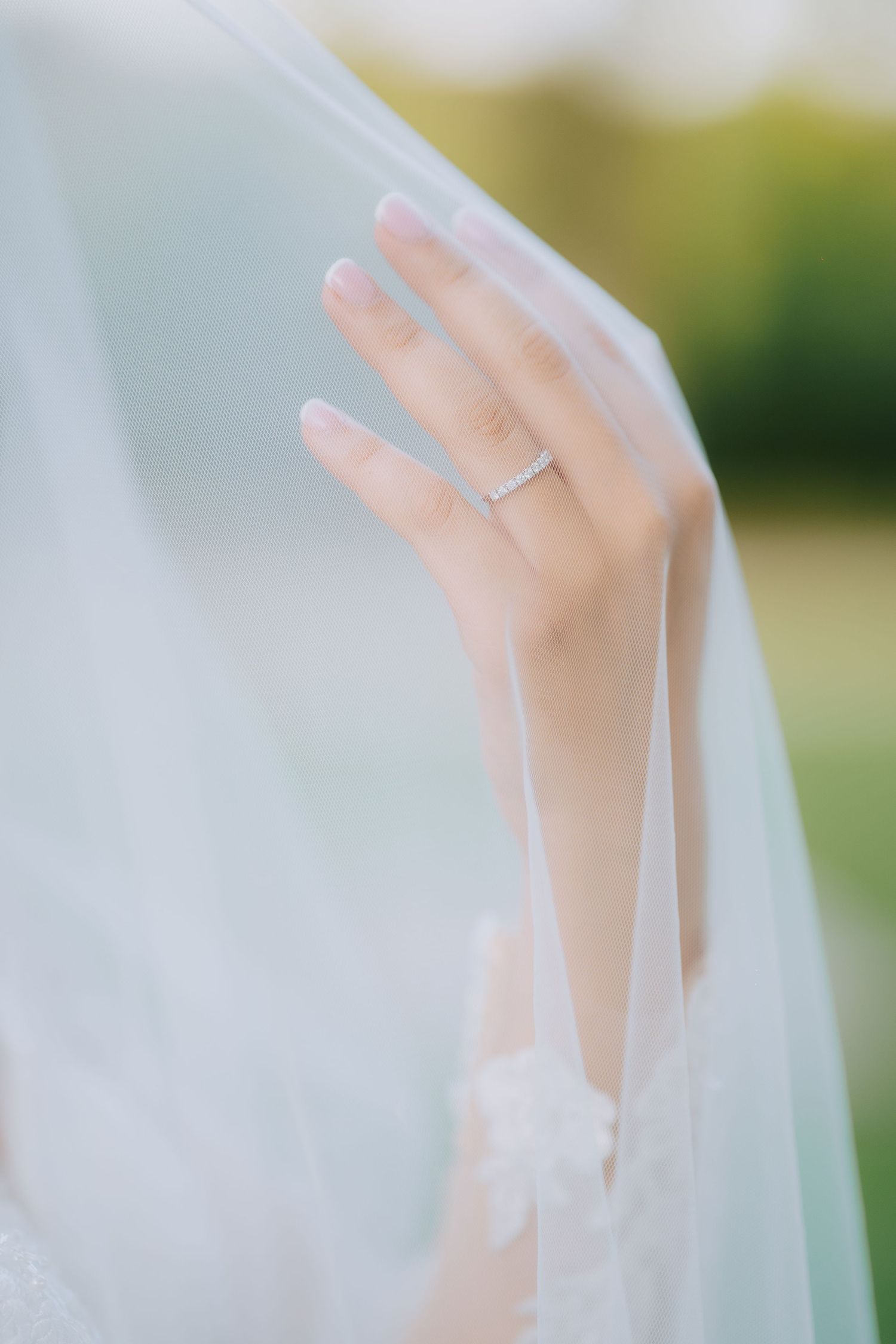 Close-up view of a delicate hand with engagement ring showing through white tulle wedding veil in soft light.
