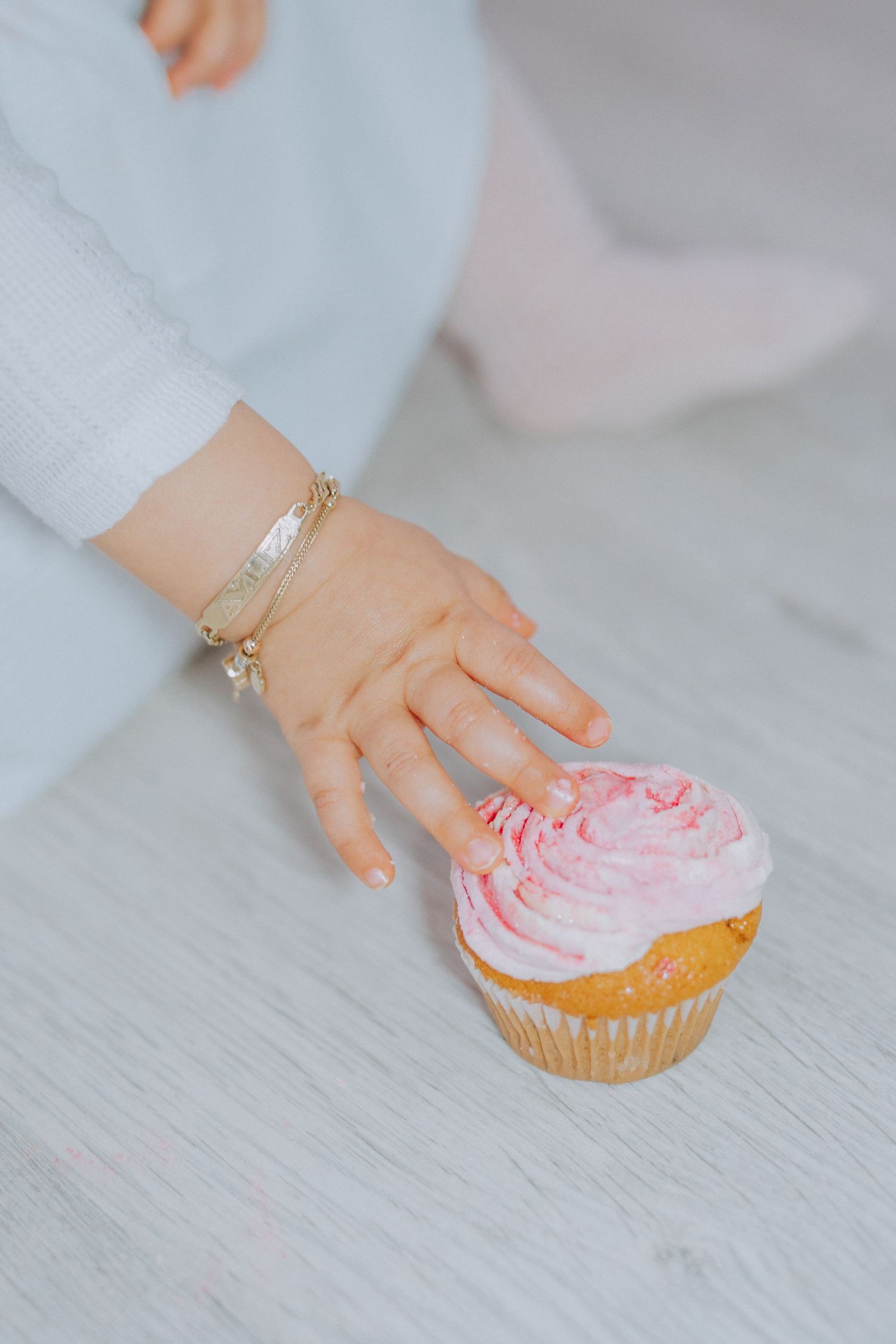 Small hand reaching for pink-frosted cupcake with gold bracelet detail.