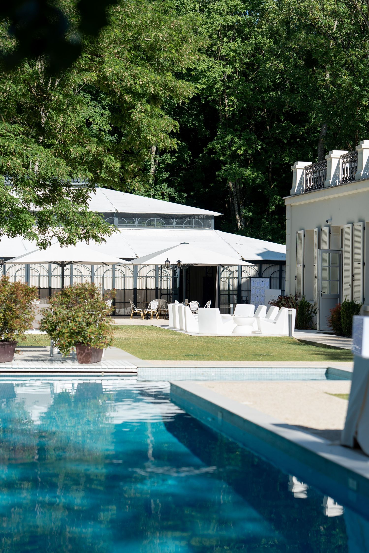 A modern rectangular swimming pool with crystal blue water surrounded by white buildings and lush green trees.