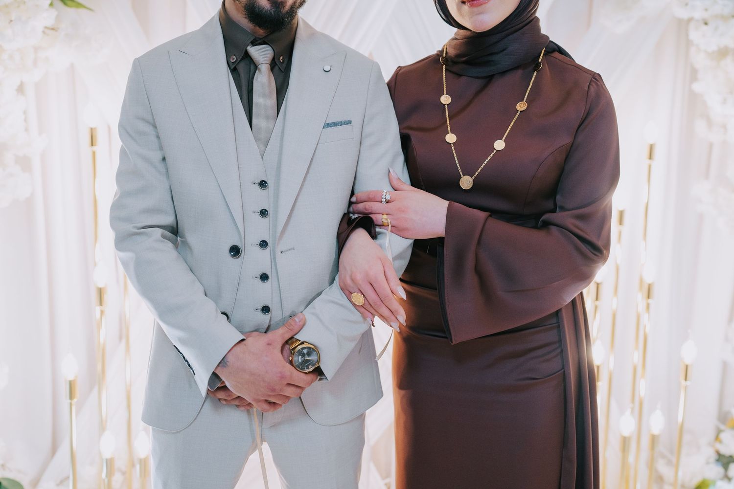 Couple in formal wear standing together, wearing grey suit and brown dress with accessories.