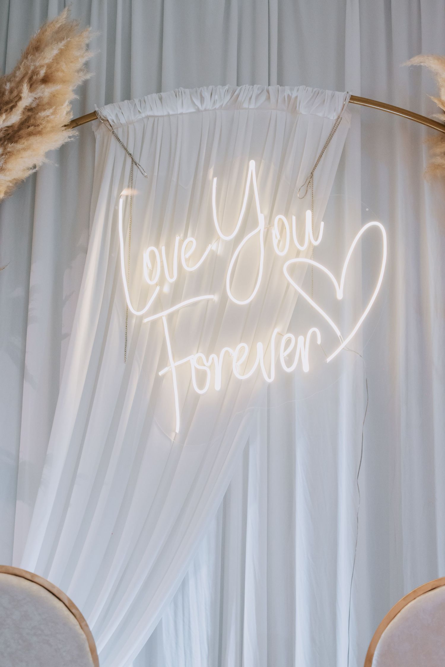 Neon 'Love You Forever' sign with heart on white draped backdrop with pampas grass.