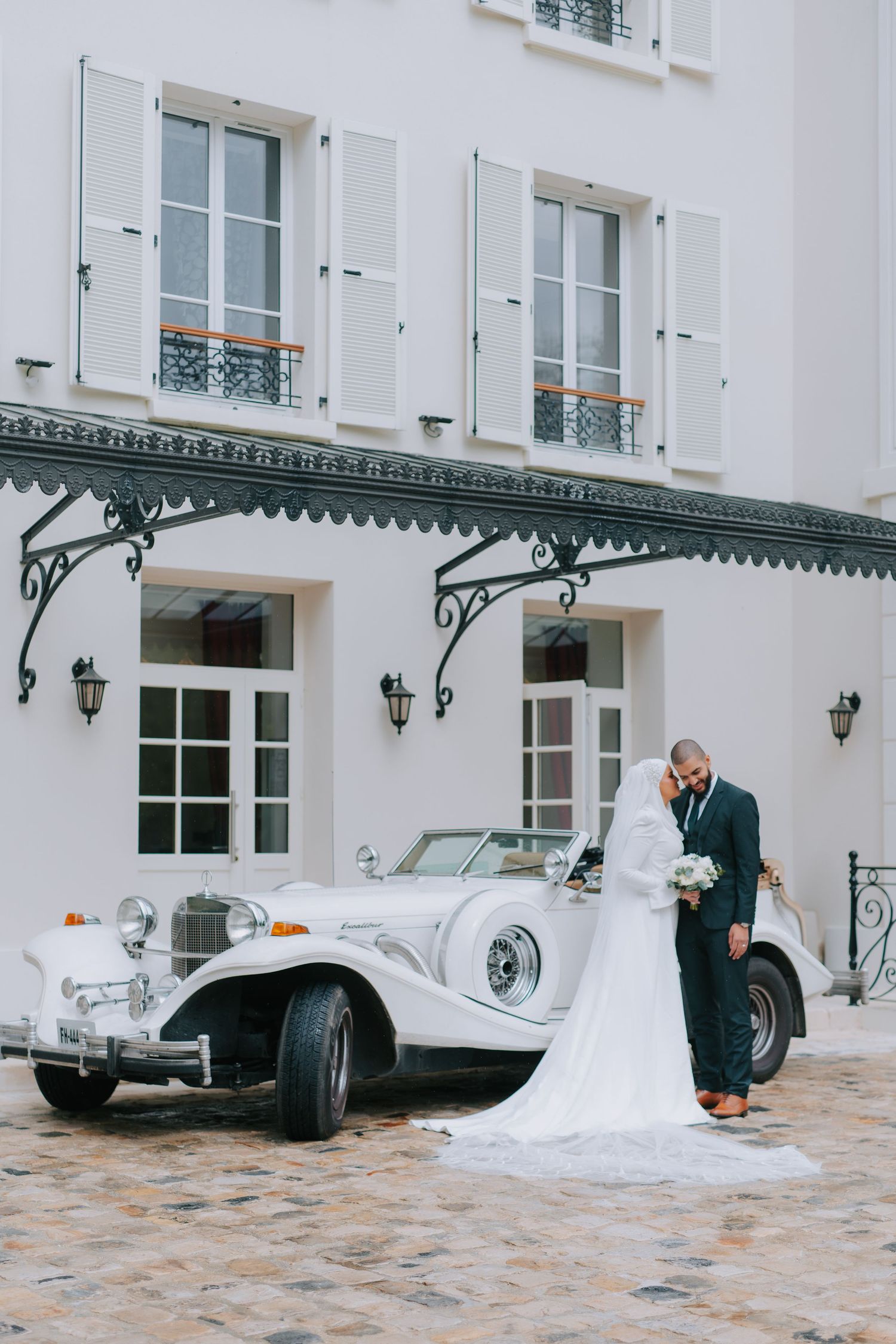 Vintage white luxury car parked in front of elegant white building with decorative metal awning on cobblestone courtyard.
