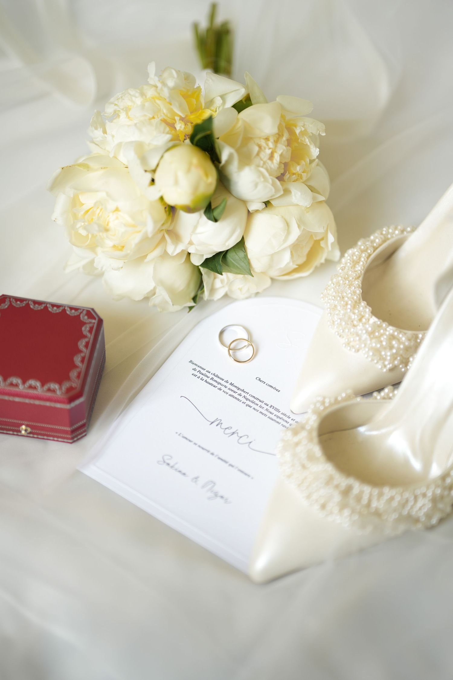 A white peony bouquet with wedding invitation, pearl-adorned heels and red jewelry box on white fabric.