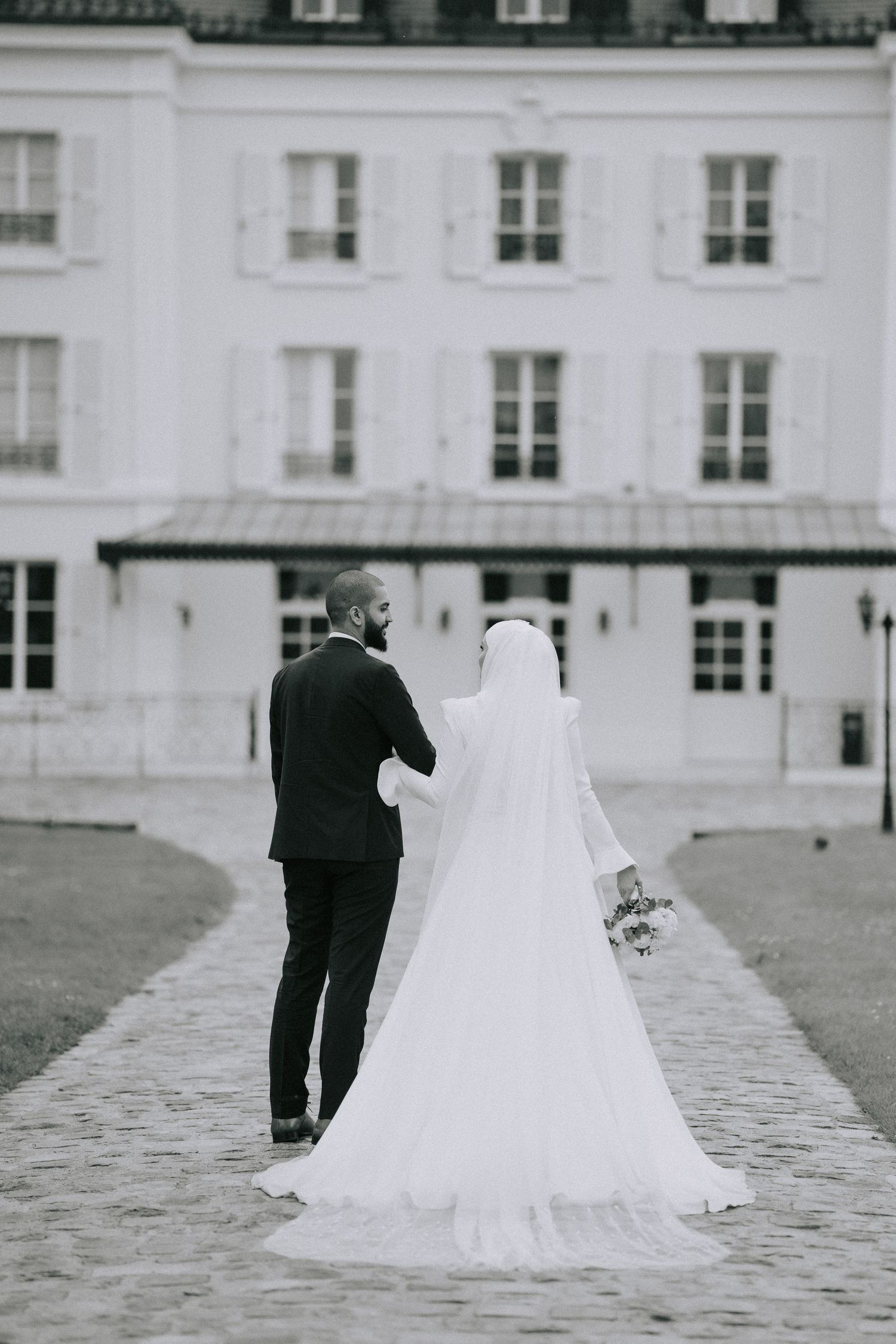 A black and white wedding photo shows a couple walking away from the camera toward a grand white building.