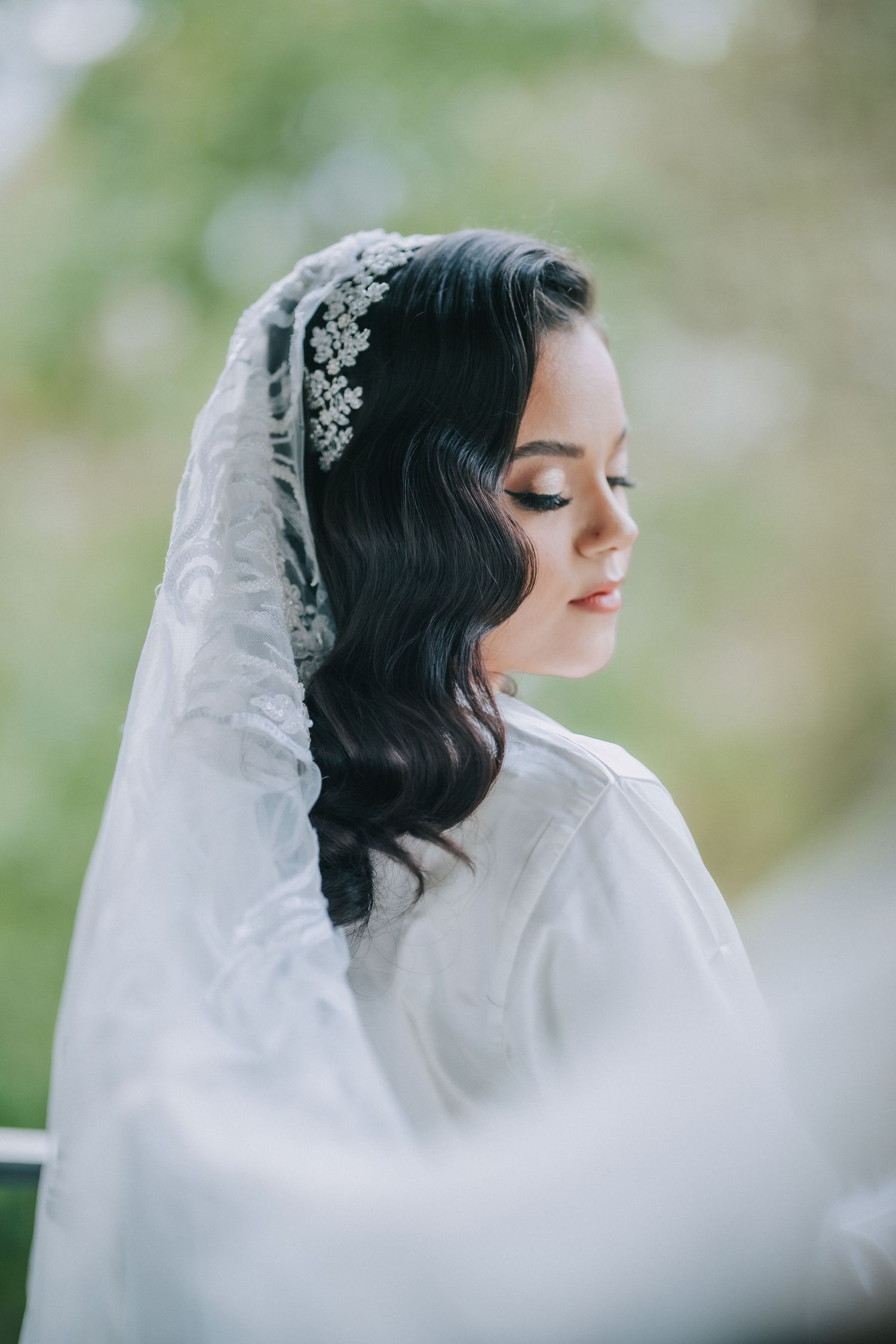 A bride in a white veil and delicate crystal hair accessory poses with eyes closed in a soft, ethereal portrait.