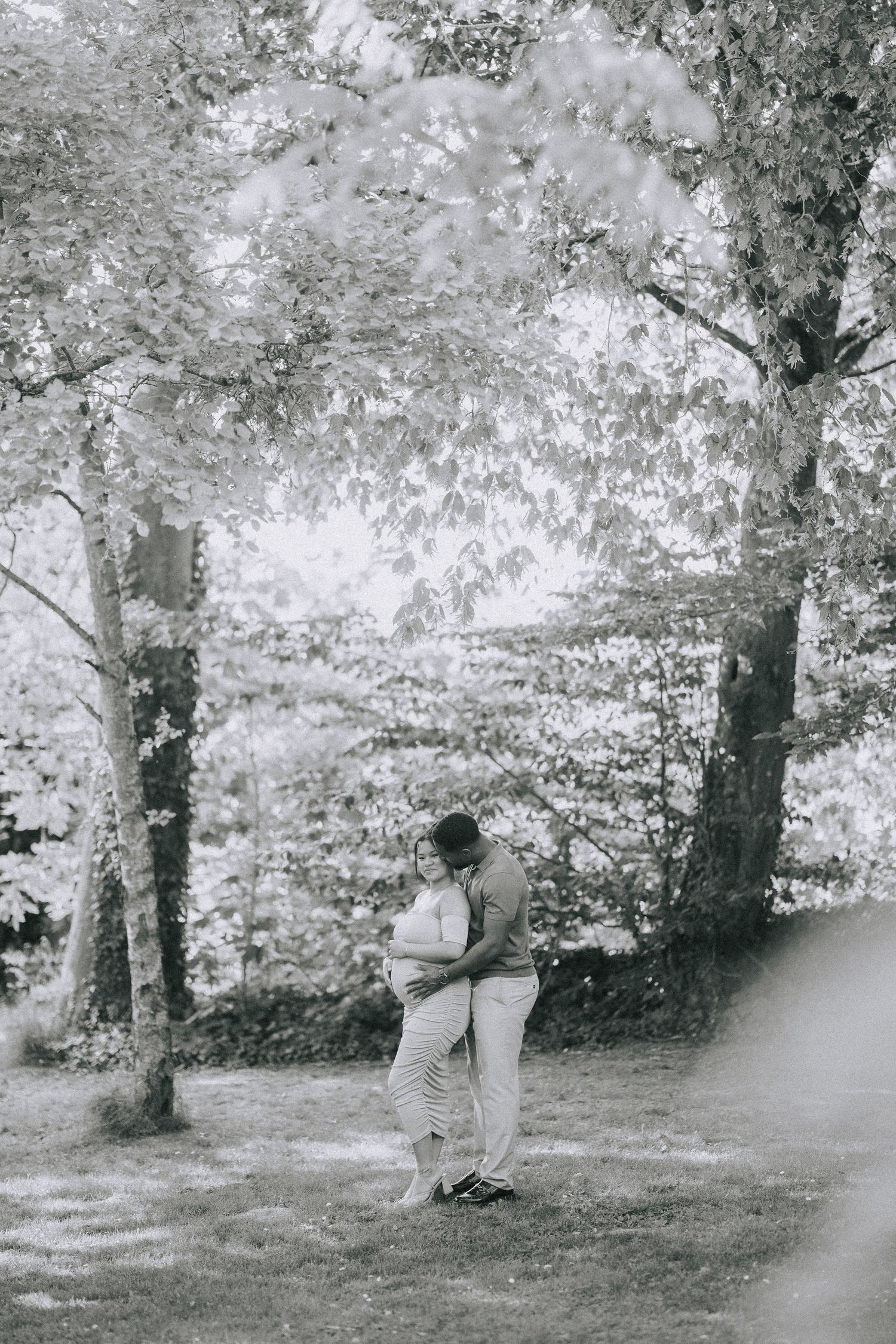 Black and white romantic photograph showing a couple embracing under leafy trees in a dreamy forest setting.