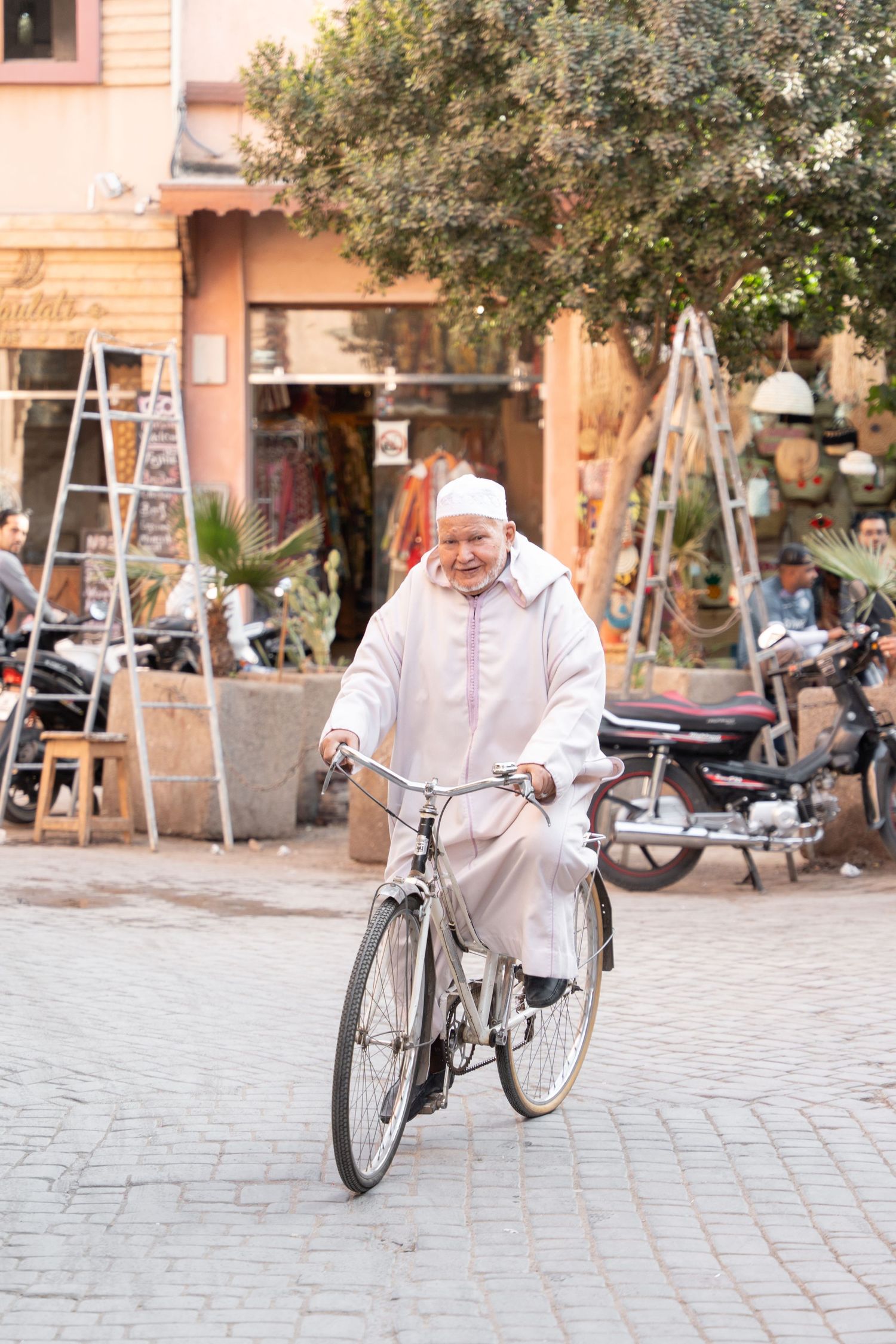 Person in traditional white dress riding bicycle on cobblestone street in Morocco.